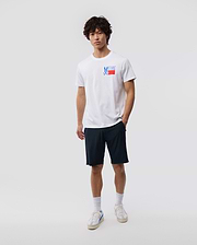 A young man with curly hair stands against a plain background, wearing a white MENS TEXAS TEE - B6U553W1PC featuring the Psycho Bunny logo graphic, navy shorts, white socks, and sneakers.