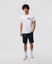 A young man with curly hair stands against a plain background, wearing a white MENS TEXAS TEE - B6U553W1PC featuring the Psycho Bunny logo graphic, navy shorts, white socks, and sneakers.