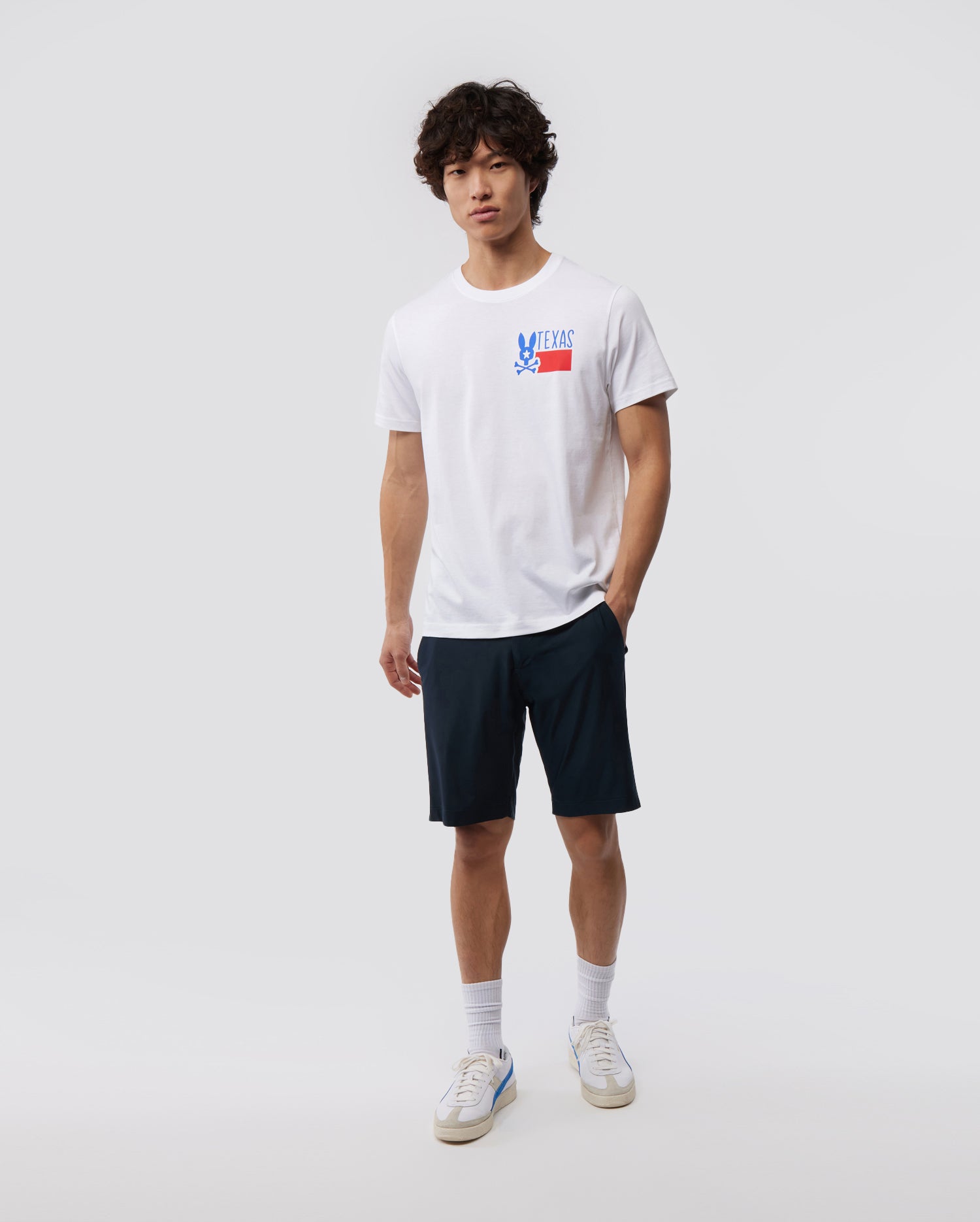 A young man with curly hair stands against a plain background, wearing a white MENS TEXAS TEE - B6U553W1PC featuring the Psycho Bunny logo graphic, navy shorts, white socks, and sneakers.