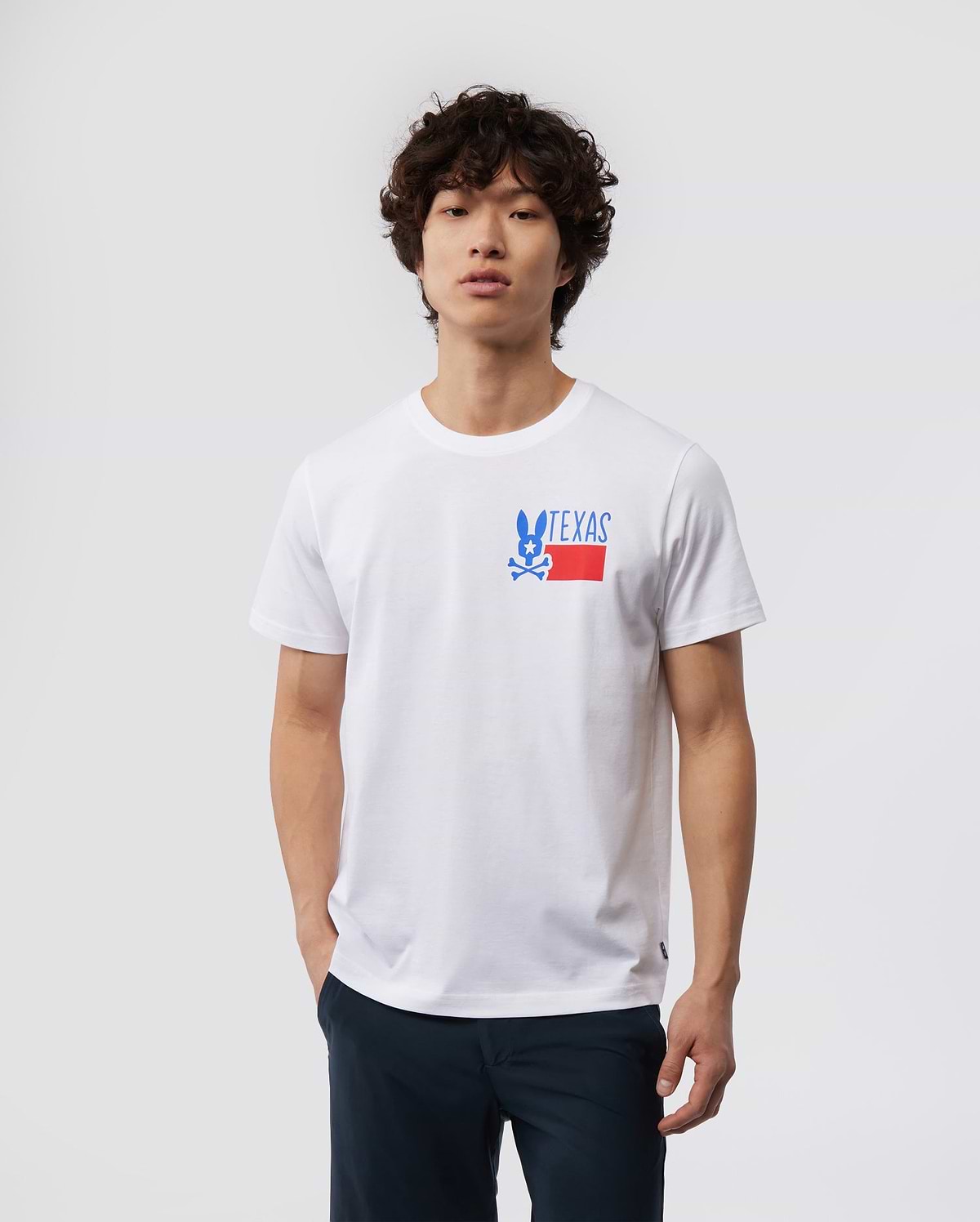 A young man with curly hair wearing a white MENS TEXAS TEE - B6U553W1PC shirt with a "Psycho Bunny" logo on the chest, paired with dark pants, standing against a plain background.