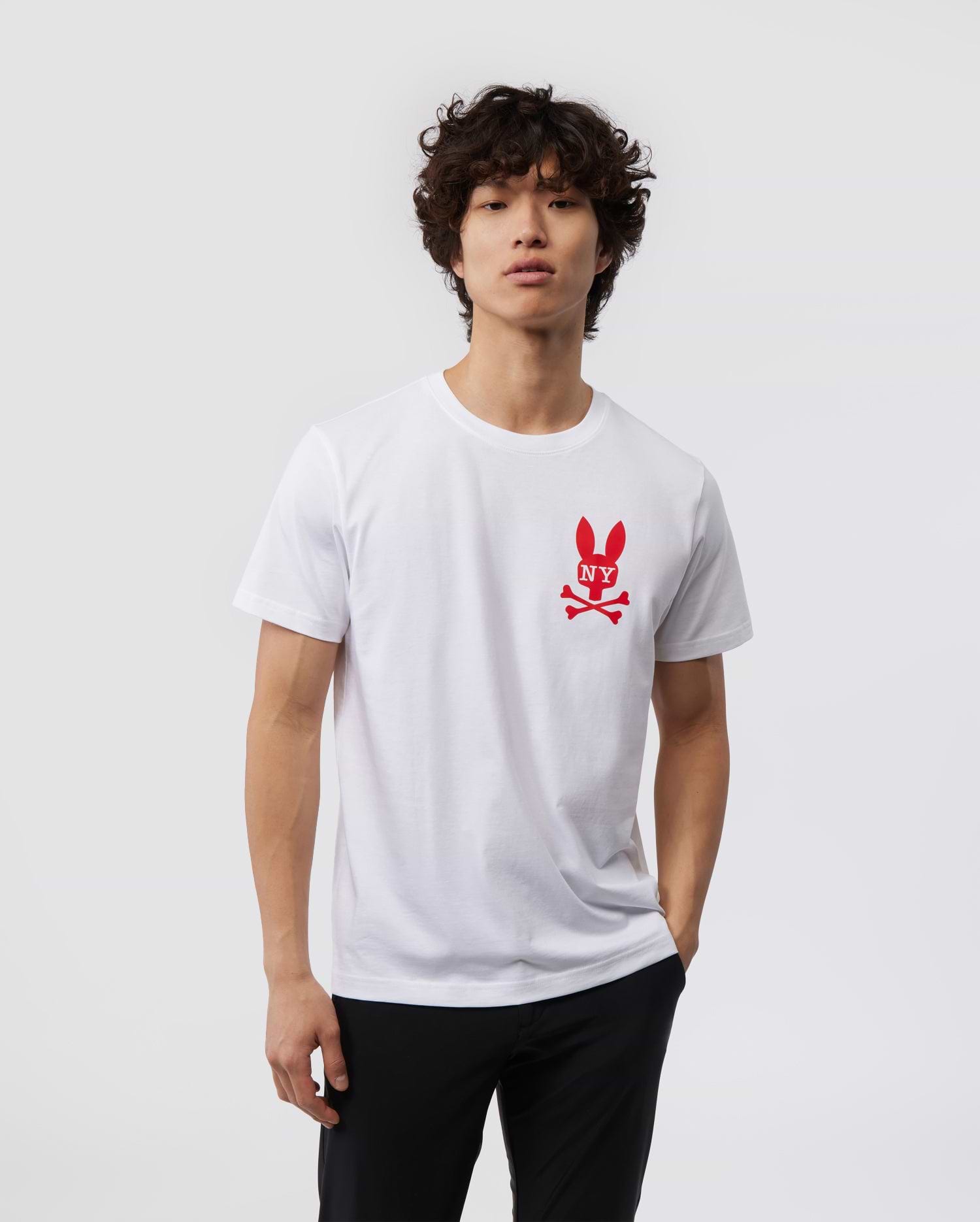 A person with short curly hair is wearing a white Pima cotton jersey T-shirt featuring a red bunny logo with crossed bones beneath it, the MENS NEW YORK TEE - B6U554W1PC from Psycho Bunny. The person is also wearing dark pants and standing against a plain light gray background.
