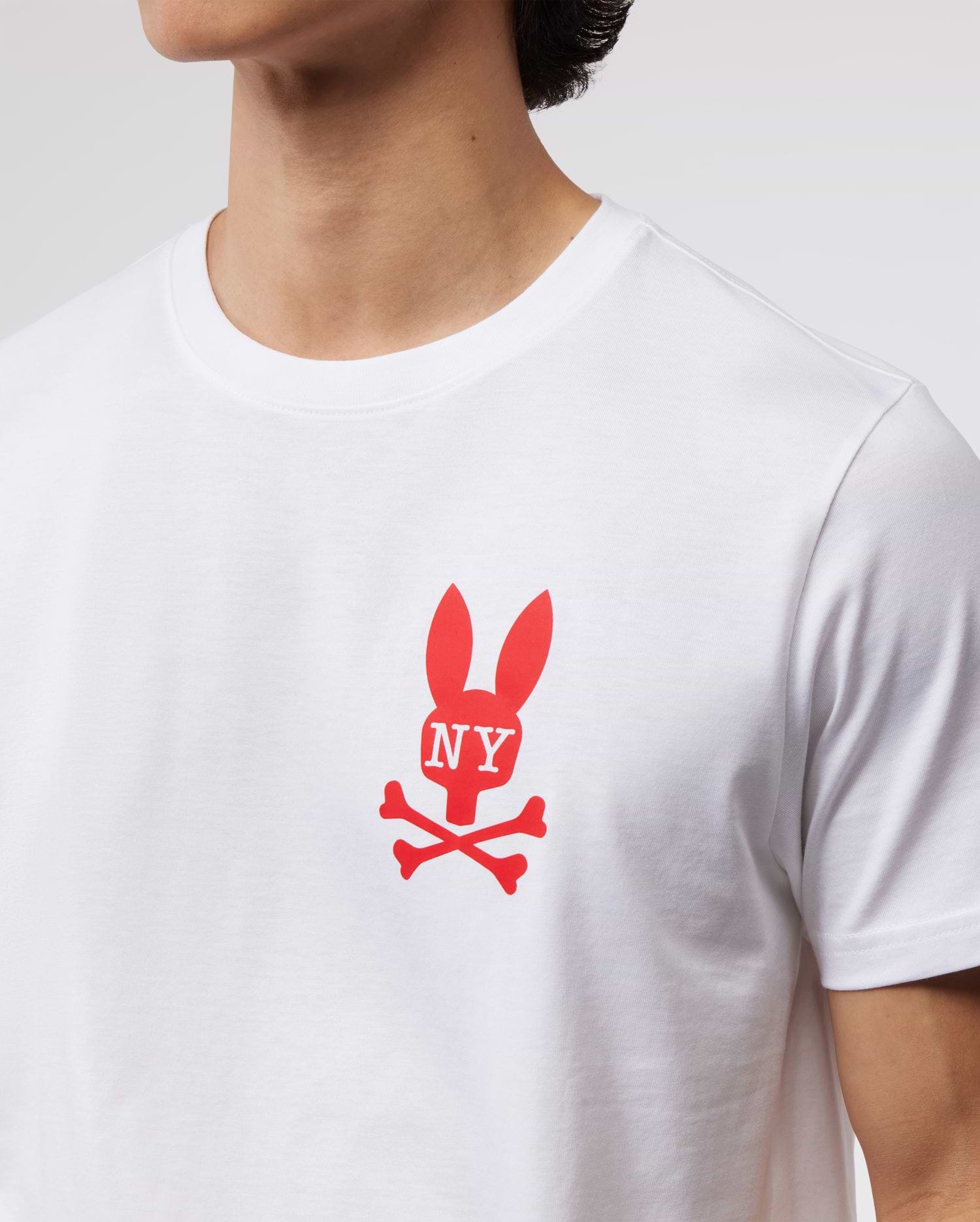 A person wearing a white Psycho Bunny MENS NEW YORK TEE - B6U554W1PC made of soft Pima cotton jersey. The red graphic on the chest features a bunny logo with ears shaped like the letters "NY" and crossed bones underneath. The photo shows the chest and neck area, but the person's face is not visible.