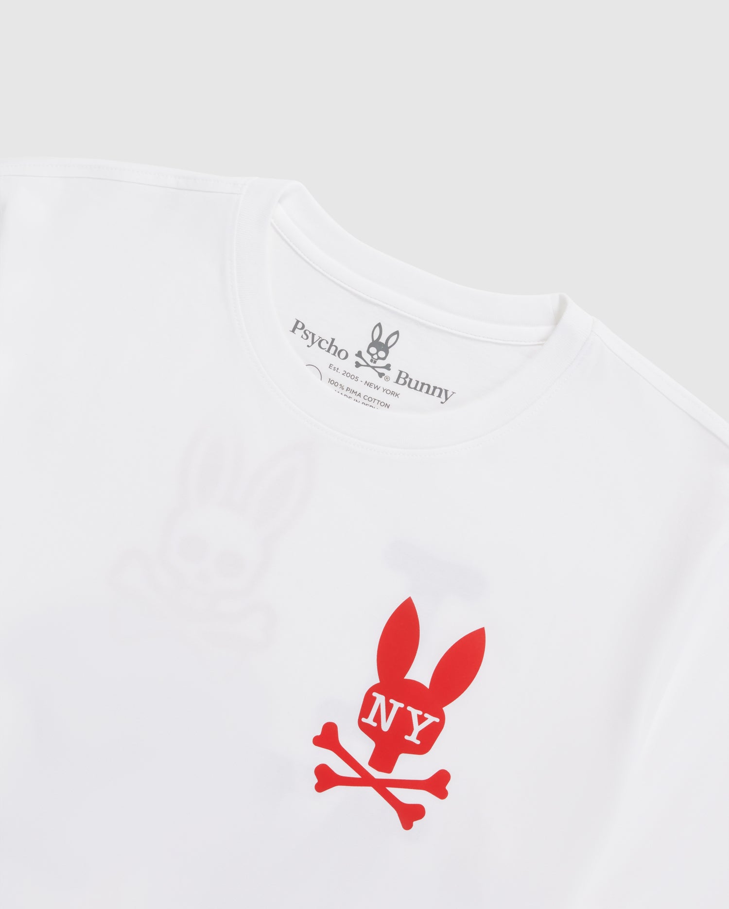 The MENS NEW YORK TEE - B6U554W1PC featuring a red bunny skull and crossbones logo with "NY" written on the bunny's head. Crafted from Pima cotton jersey, the shirt’s brand name, "Psycho Bunny," is printed on the inside neckline tag in black text, accompanied by a small bunny logo above it.