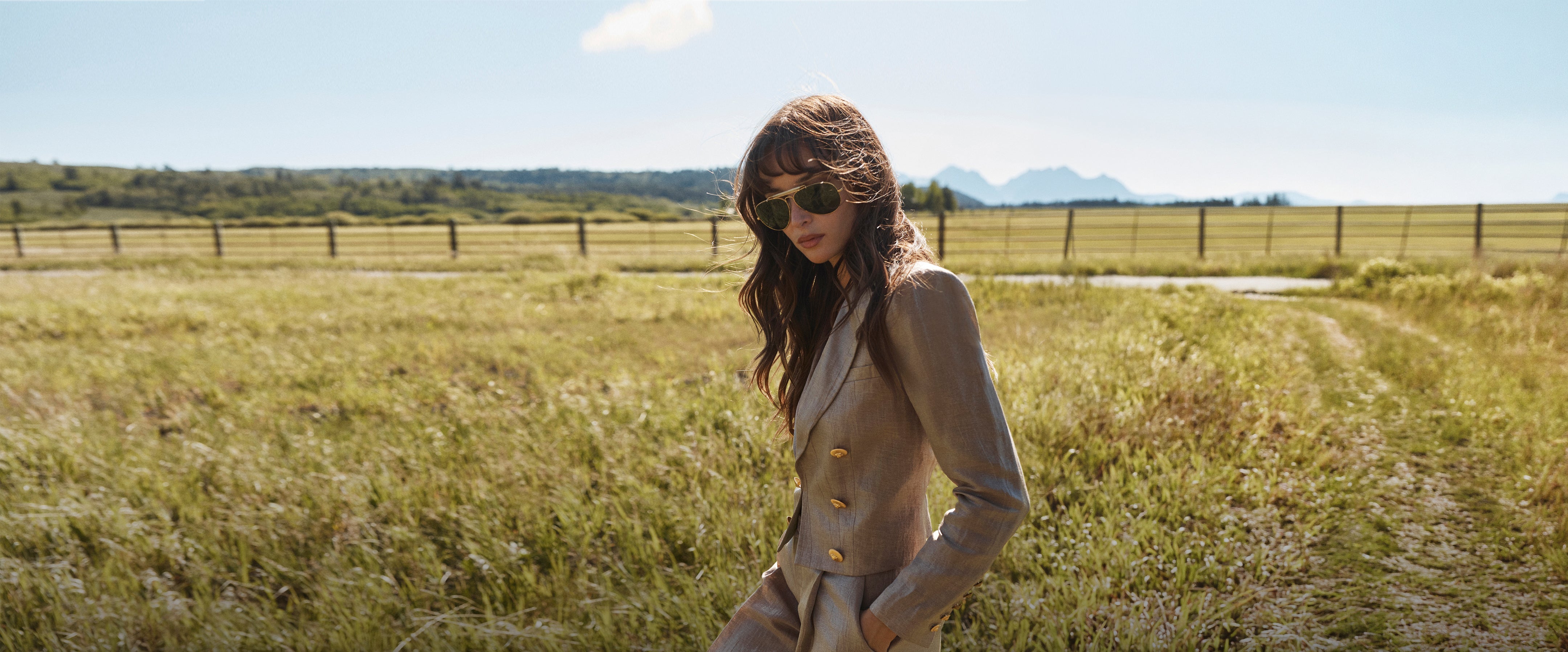 A woman in sunglasses and a plaid suit stands by a wooden fence in a grassy field, with a road and trees in the background.