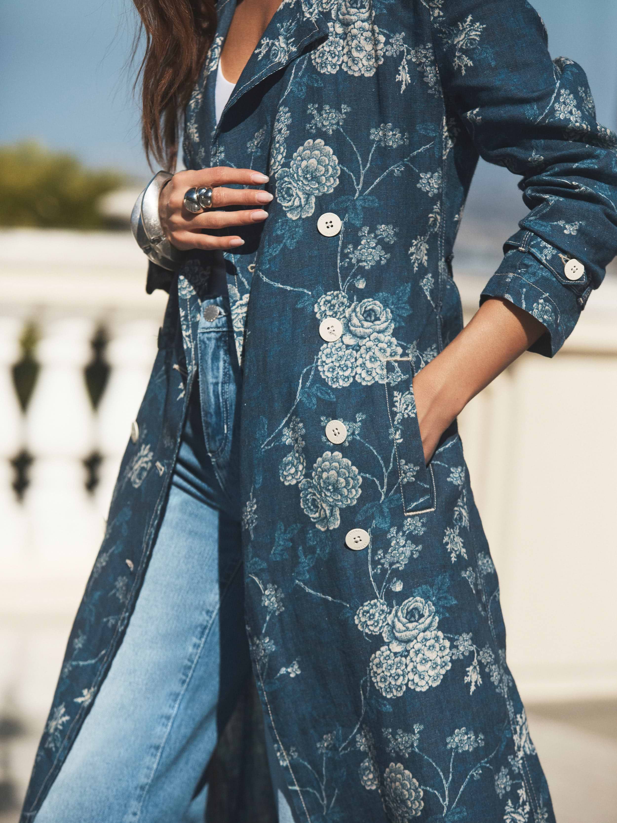 A person stands outdoors wearing a blue floral patterned trench coat over blue jeans, with one hand in the coat pocket and the other adjusting the lapel. The background is bright and blurred.