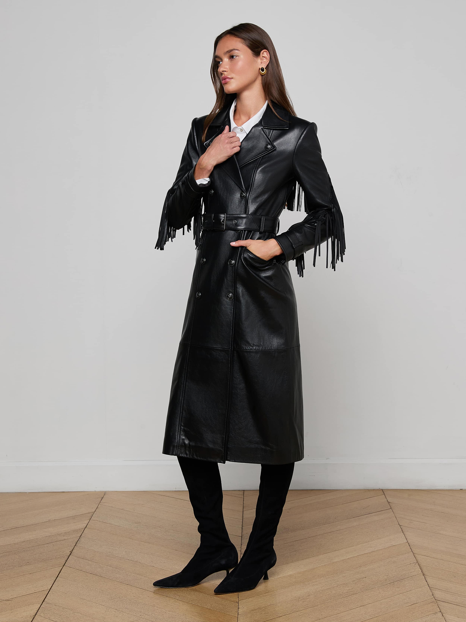 Wearing the Celina Fringed Leather Trench with its dramatic fringed sleeve trim, black boots, and a white shirt, the woman stands against a plain background with one hand in her pocket.