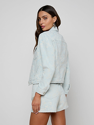 A woman models the Maven Jacket, a structured-fit, light blue denim piece paired with matching shorts. She stands sideways and glances over her shoulder against a plain, light background.