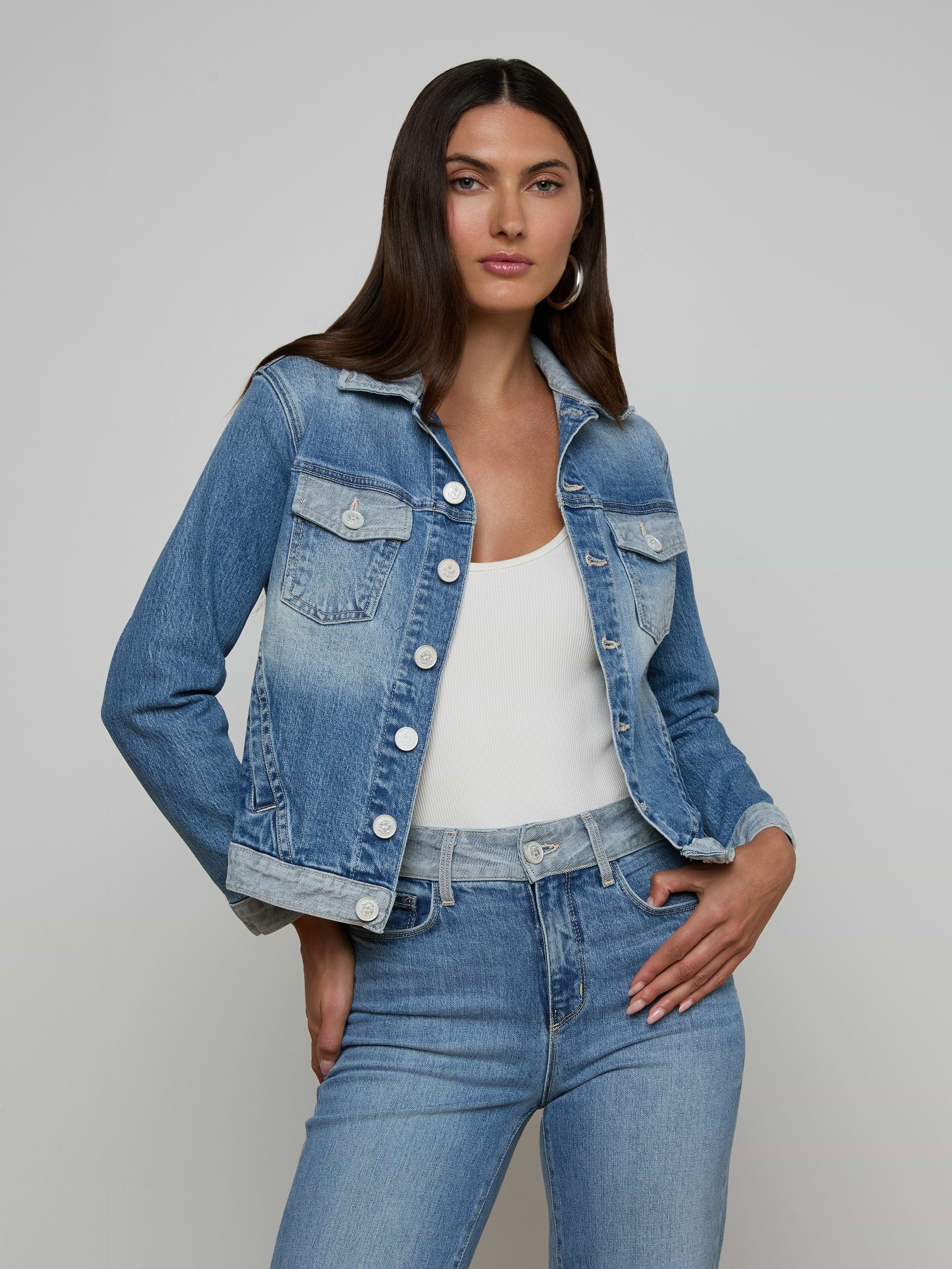 A woman stands against a plain light background with one hand on her hip, wearing the Maven Jean Jacket—a light-wash, three-quarter sleeve jean jacket—over a white top and blue jeans.