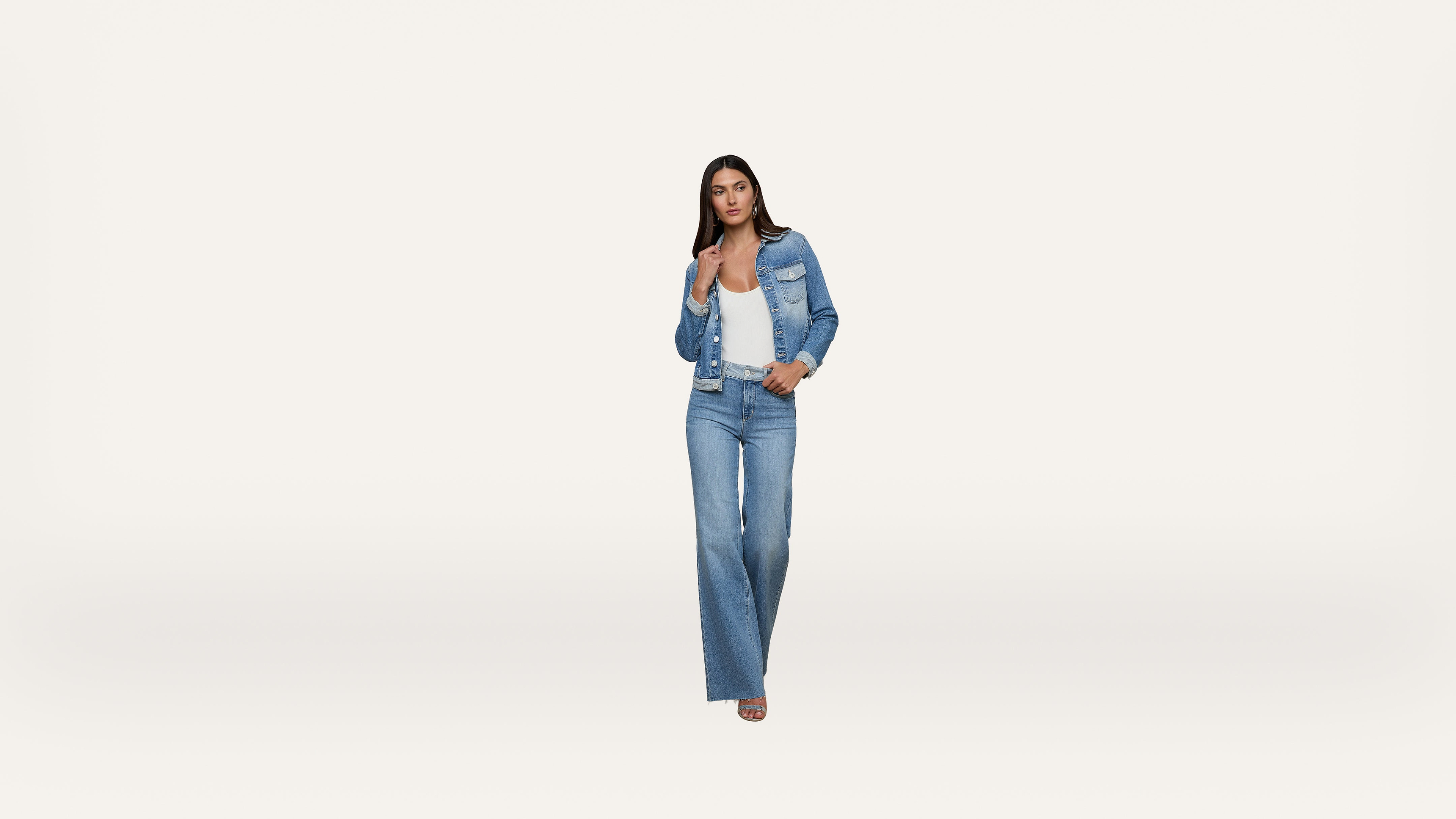 A woman stands facing forward, wearing a blue denim jacket and flared jeans with a white top and sandals, against a plain white background.