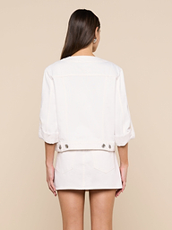 A woman stands facing away, wearing the Anouk Jean Jacket with rolled sleeves and a matching skirt. Her long brown hair is down, and her hands rest by her sides against a plain background.