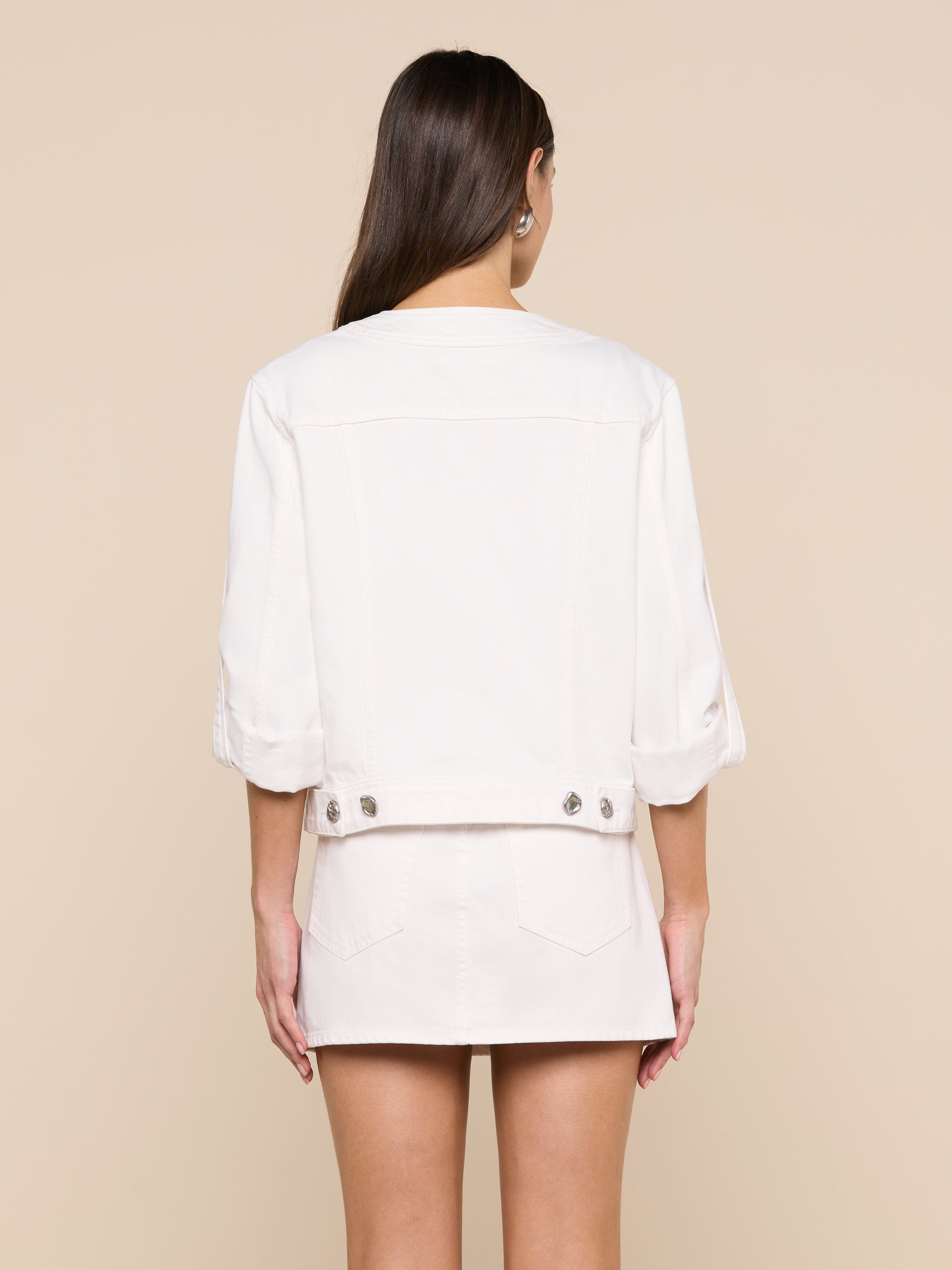 A woman stands facing away, wearing the Anouk Jean Jacket with rolled sleeves and a matching skirt. Her long brown hair is down, and her hands rest by her sides against a plain background.