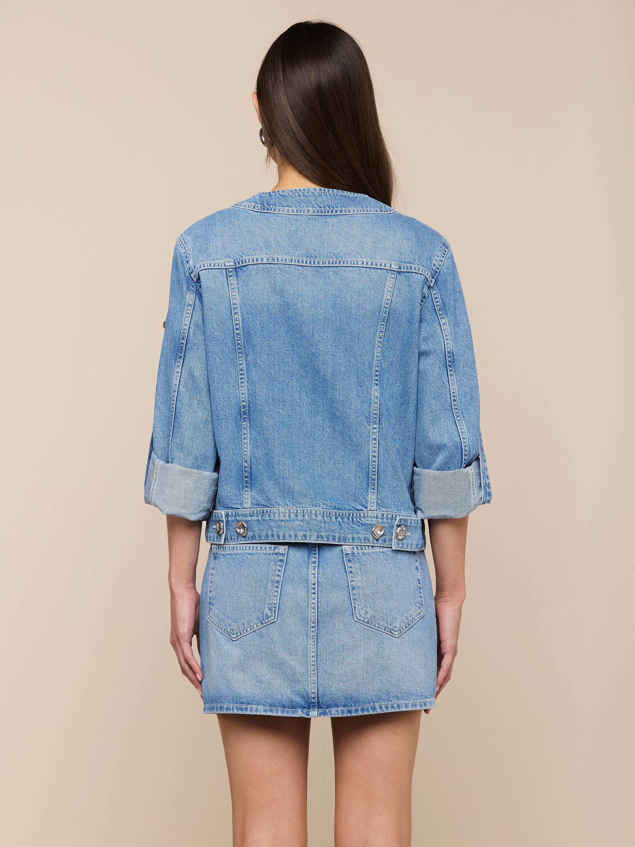 A woman with long brown hair is shown from the back against a plain light background, wearing the Anouk Jean Jacket—a relaxed fit, collarless denim jacket with rolled sleeves—paired with a matching denim skirt.