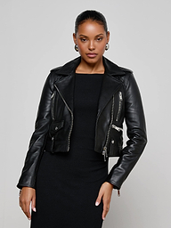 Wearing the Blakely Leather Biker Jacket over a black dress, a woman stands against a plain light background, looking confidently at the camera with one hand on her hip.