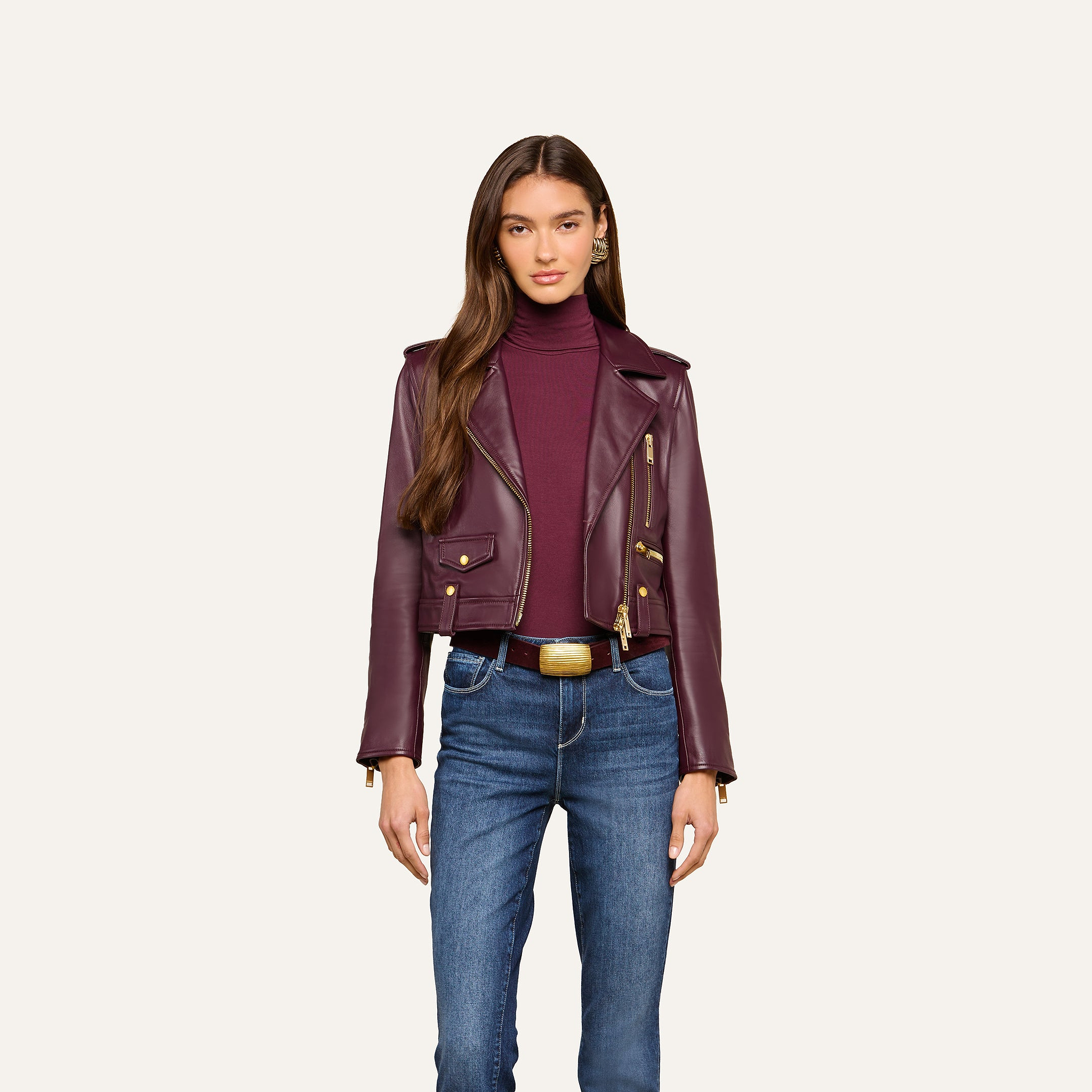 A woman stands facing forward against a plain, light background, wearing the Blakely Leather Biker Jacket in maroon lambskin over a matching turtleneck and blue jeans.