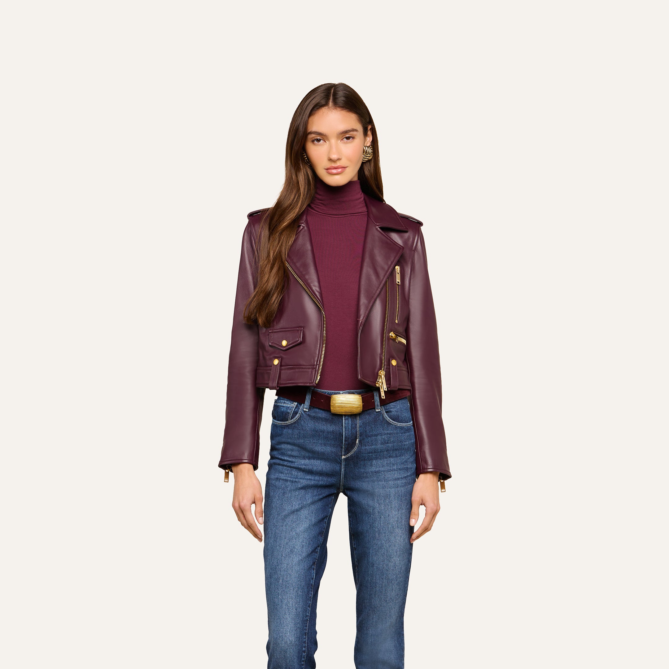 A woman stands facing forward against a plain, light background, wearing the Blakely Leather Biker Jacket in maroon lambskin over a matching turtleneck and blue jeans.