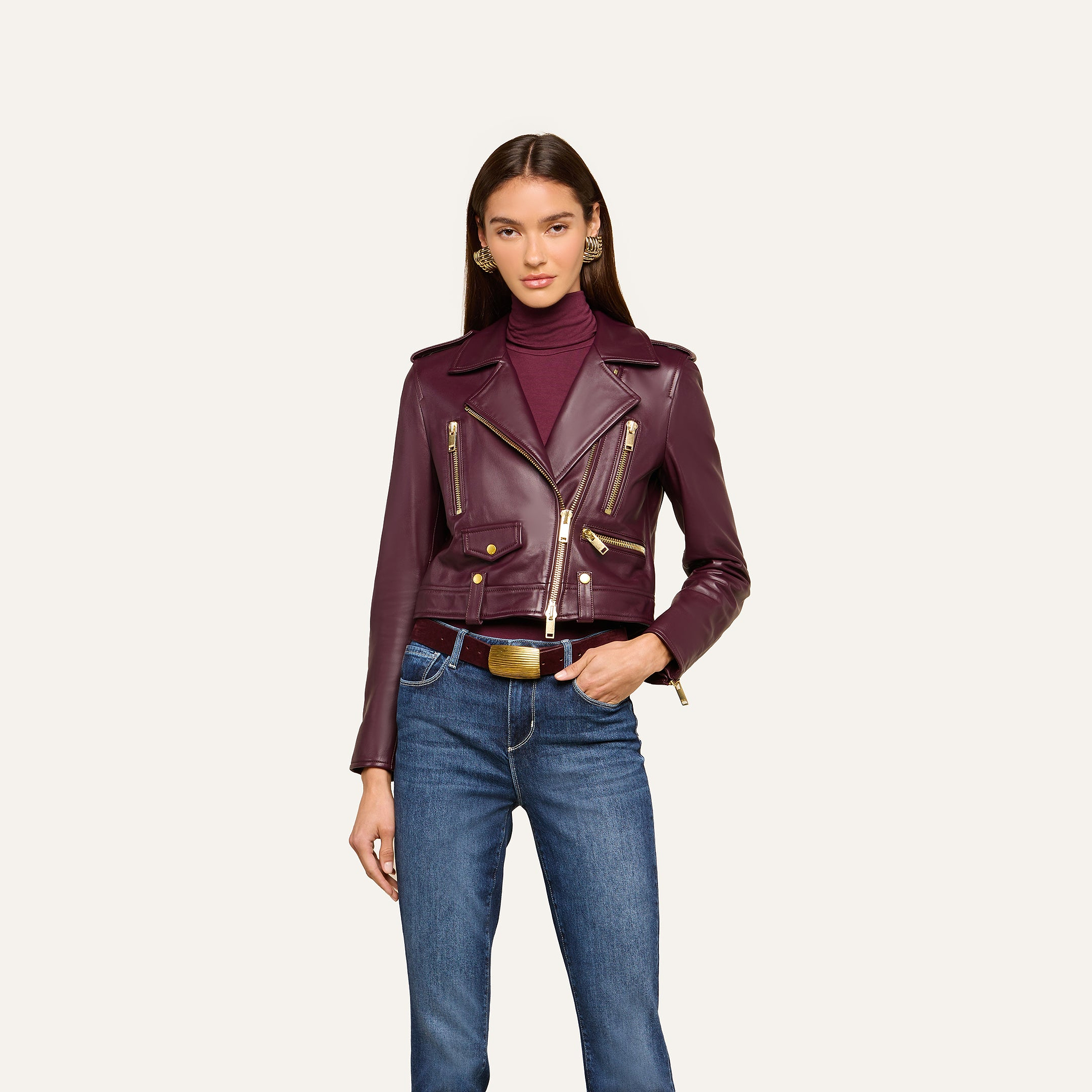A woman wears the Blakely Leather Biker Jacket with a cropped fit over a burgundy turtleneck and blue jeans. She has long straight hair and gold earrings, standing against a plain light background.