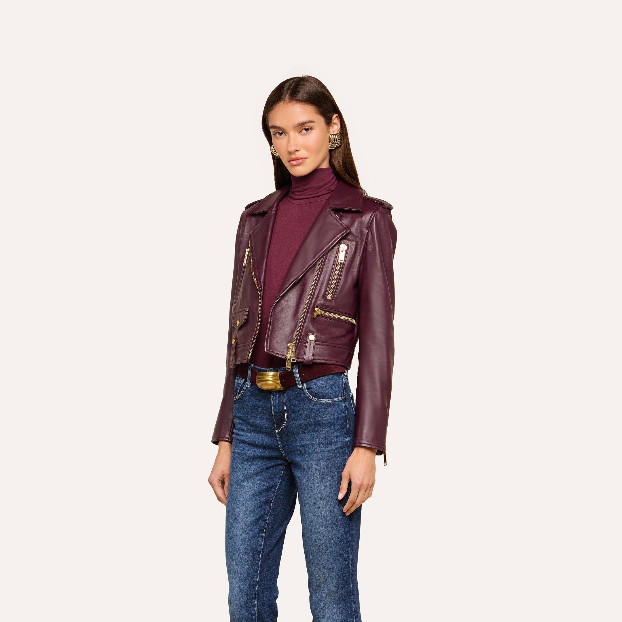 A woman wearing the Blakely Leather Biker Jacket in burgundy over a matching turtleneck and blue jeans stands against a plain light background.