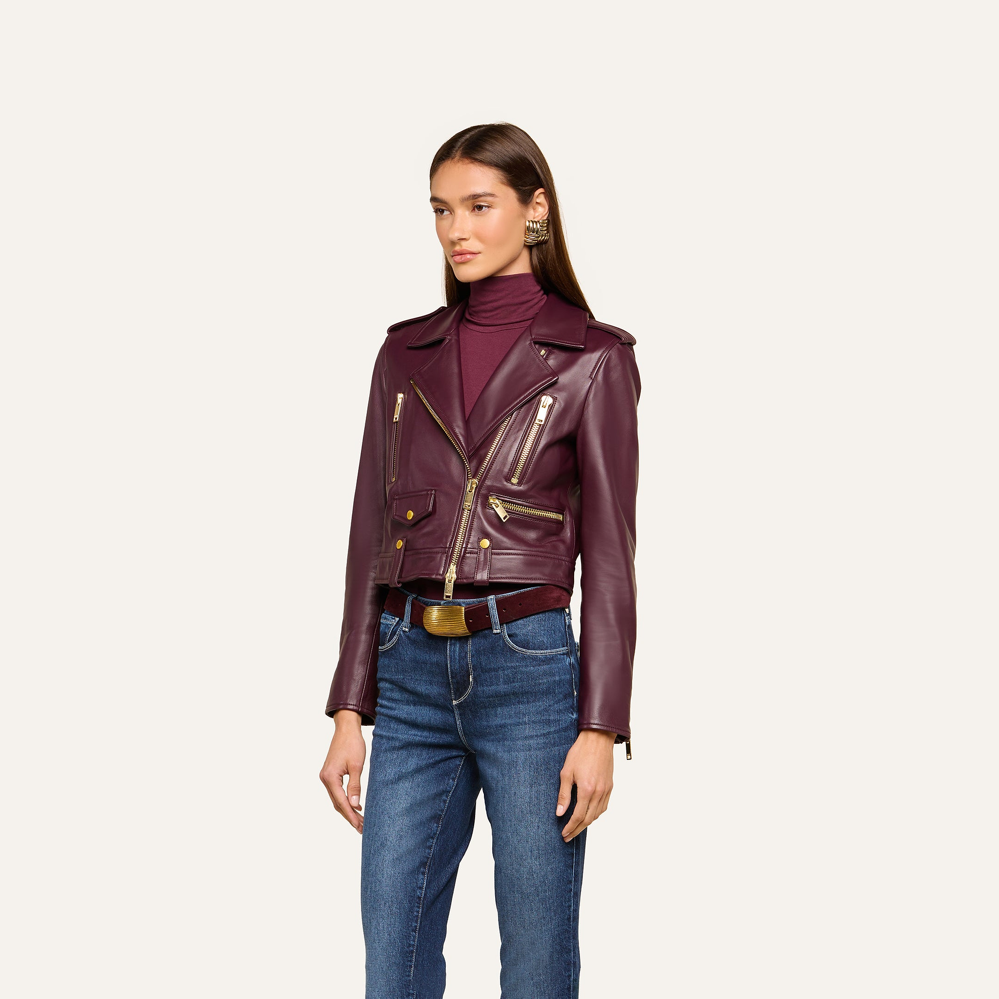 Woman wearing the Blakely Leather Biker Jacket—a maroon cropped fit with gold zippers—paired with a matching maroon turtleneck, jeans, and a brown belt, standing against a plain light background.