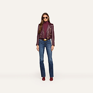 A woman stands against a plain white background, facing forward with her hands by her sides, wearing sunglasses, blue jeans, burgundy shoes, a matching turtleneck, and the Blakely Leather Biker Jacket in a cropped fit.