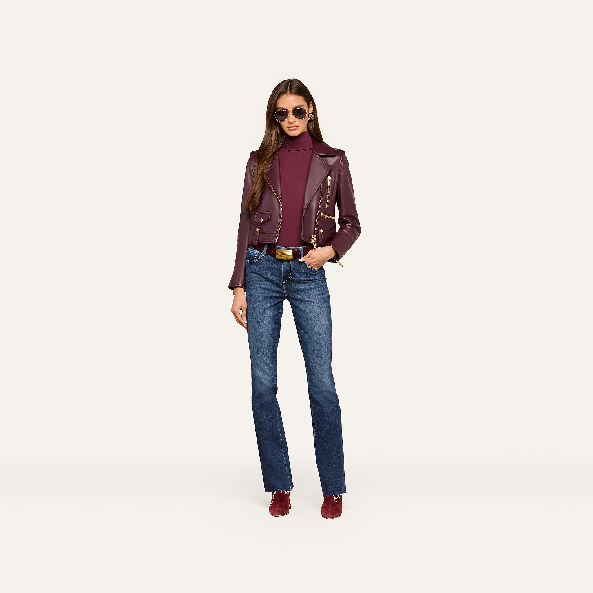 Wearing sunglasses, a woman models the Blakely Leather Biker Jacket in maroon lambskin leather over a matching maroon turtleneck, paired with belted blue jeans and maroon shoes, standing against a plain light background.