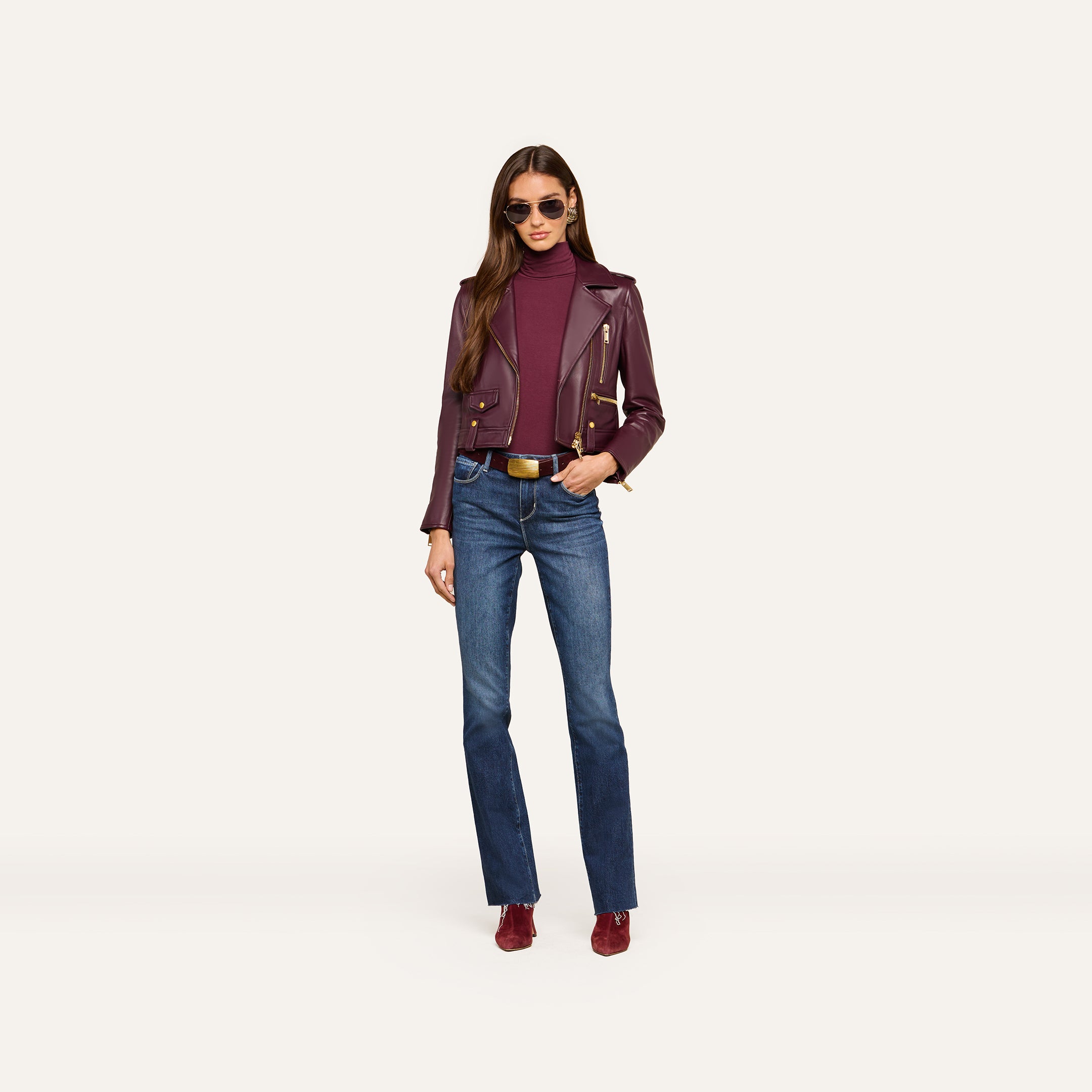 Wearing sunglasses, a woman models the Blakely Leather Biker Jacket in maroon lambskin leather over a matching maroon turtleneck, paired with belted blue jeans and maroon shoes, standing against a plain light background.