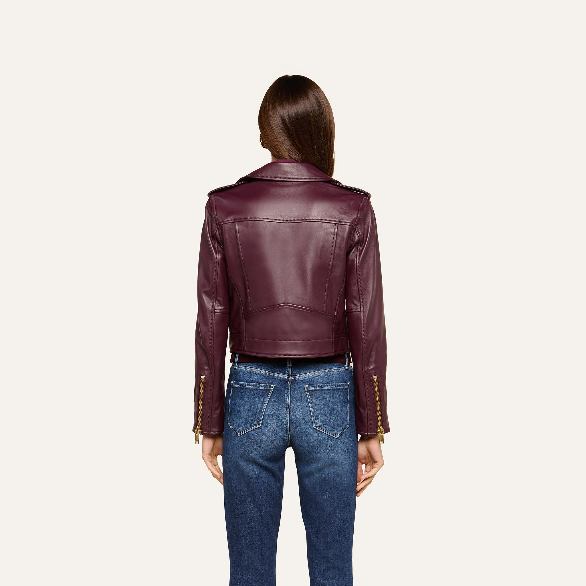 A person with long brown hair is shown from behind, wearing the Blakely Leather Biker Jacket in burgundy lambskin leather with a cropped fit and blue jeans, standing against a plain light background.