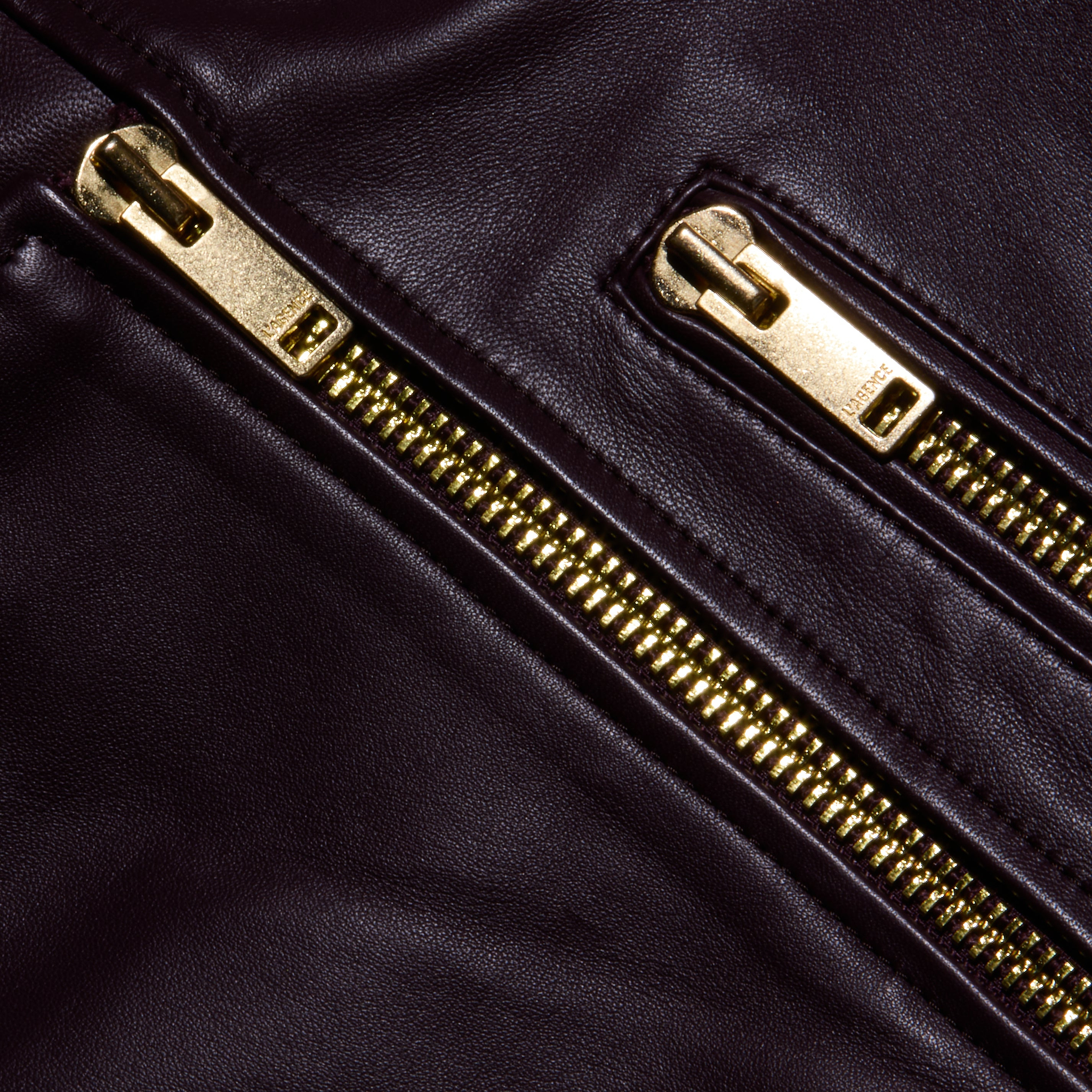 Close-up of two gold zippers on dark brown lambskin leather, highlighting the shiny metal teeth and smooth texture characteristic of the Blakely Leather Biker Jacket with its cropped fit.