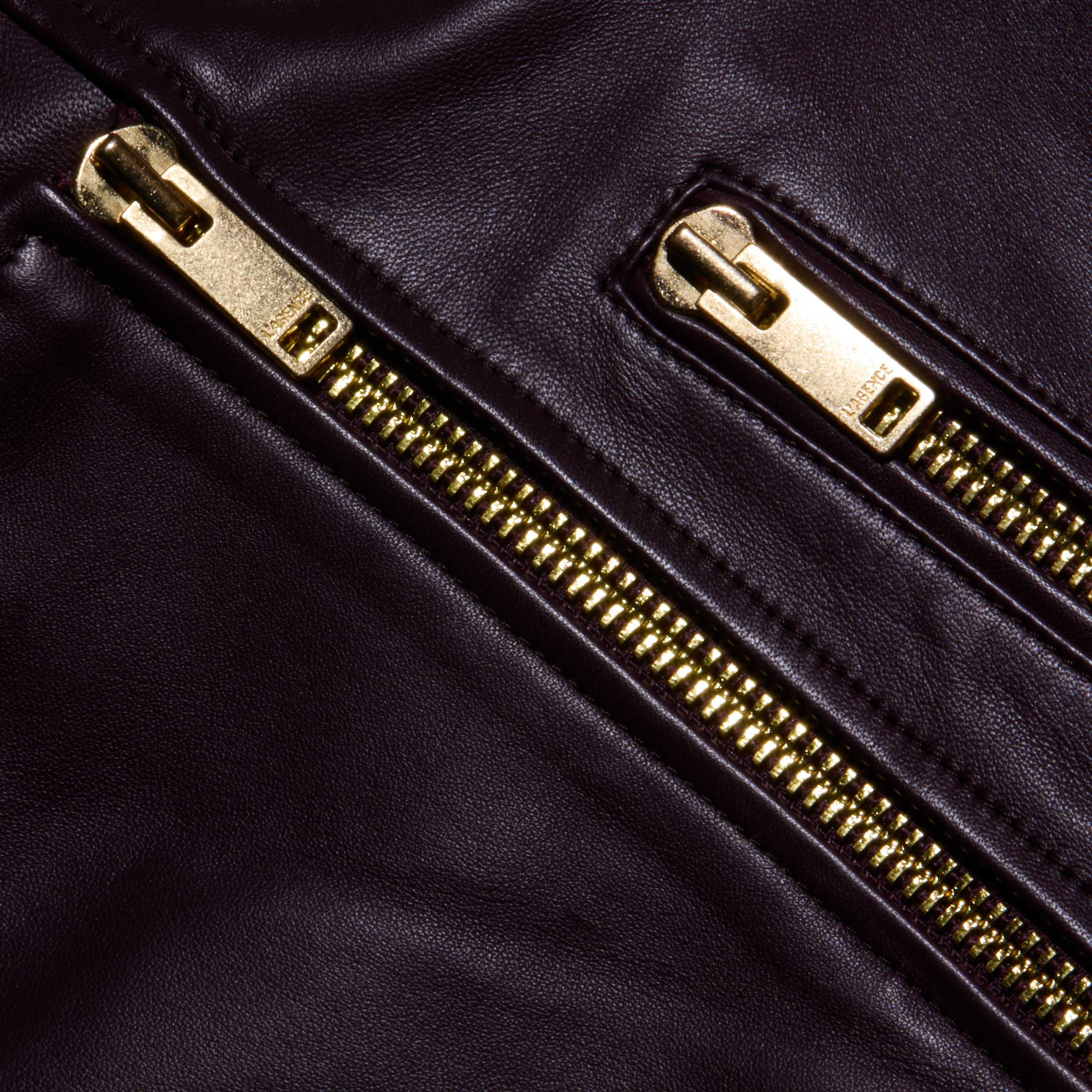 Close-up of two gold zippers on dark brown lambskin leather, highlighting the shiny metal teeth and smooth texture characteristic of the Blakely Leather Biker Jacket with its cropped fit.
