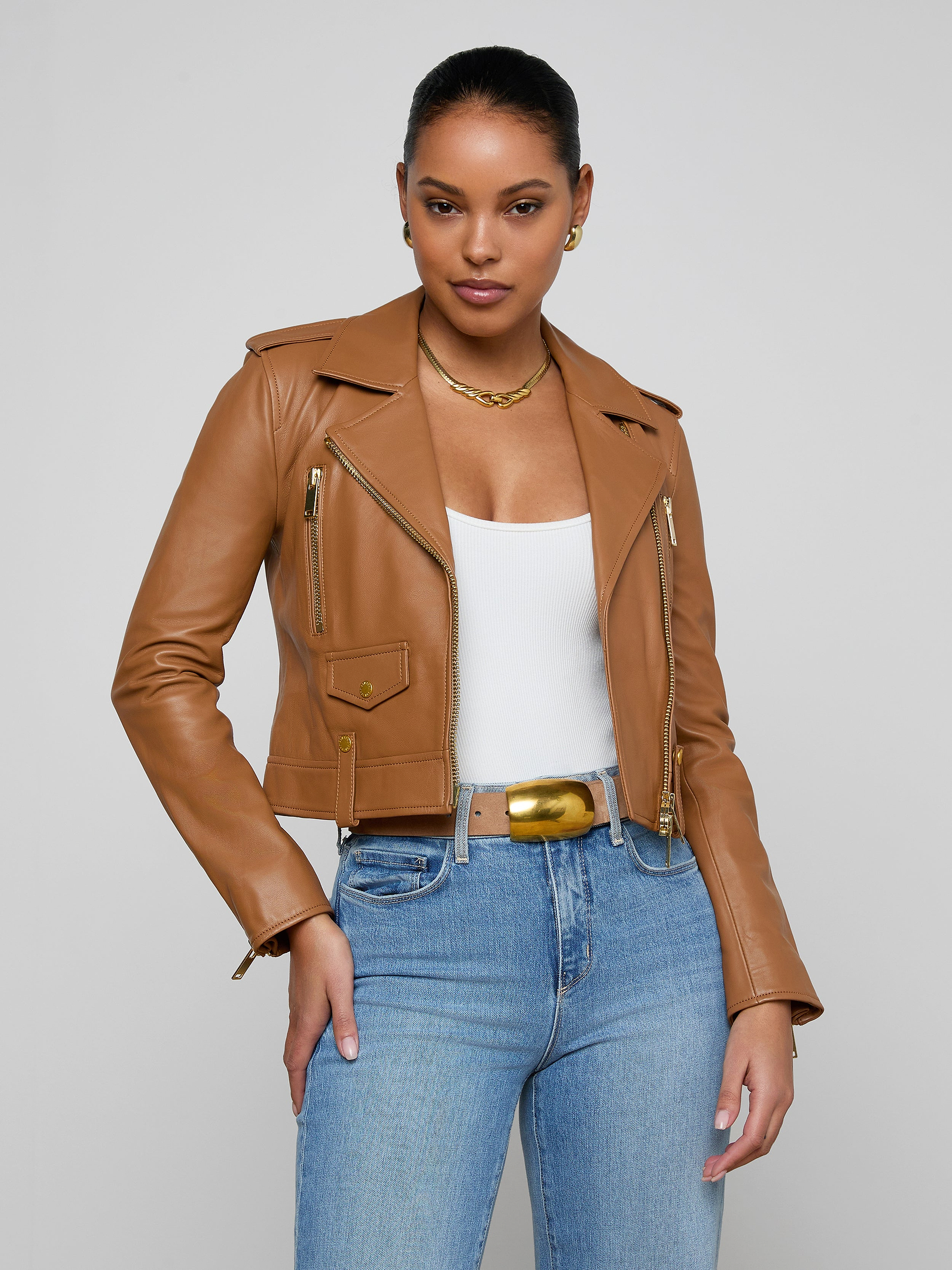 A woman wears the Blakely Leather Biker Jacket, a tan cropped motorcycle style crafted from lambskin leather, layered over a white top and blue jeans. She accessorizes with a gold necklace and belt, standing against a plain light background.