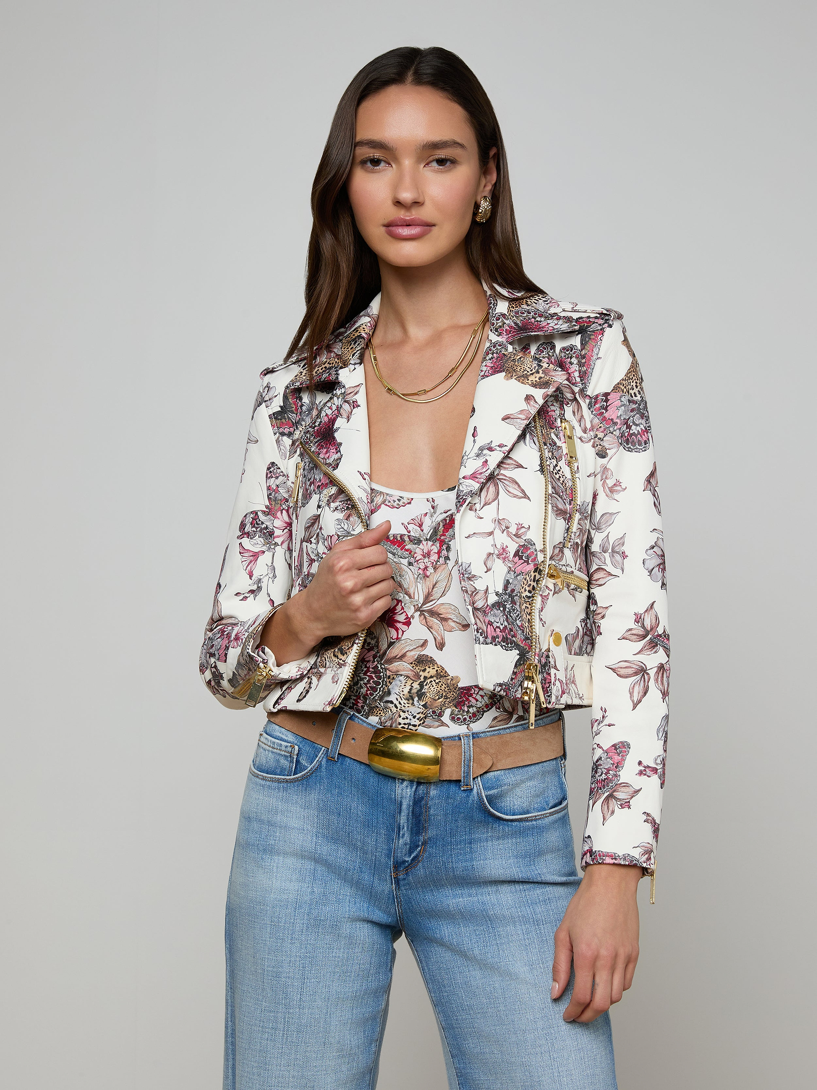 Woman wearing the Blakely Leather Biker Jacket, blue jeans, a gold belt, and layered necklaces, standing against an off-white background.
