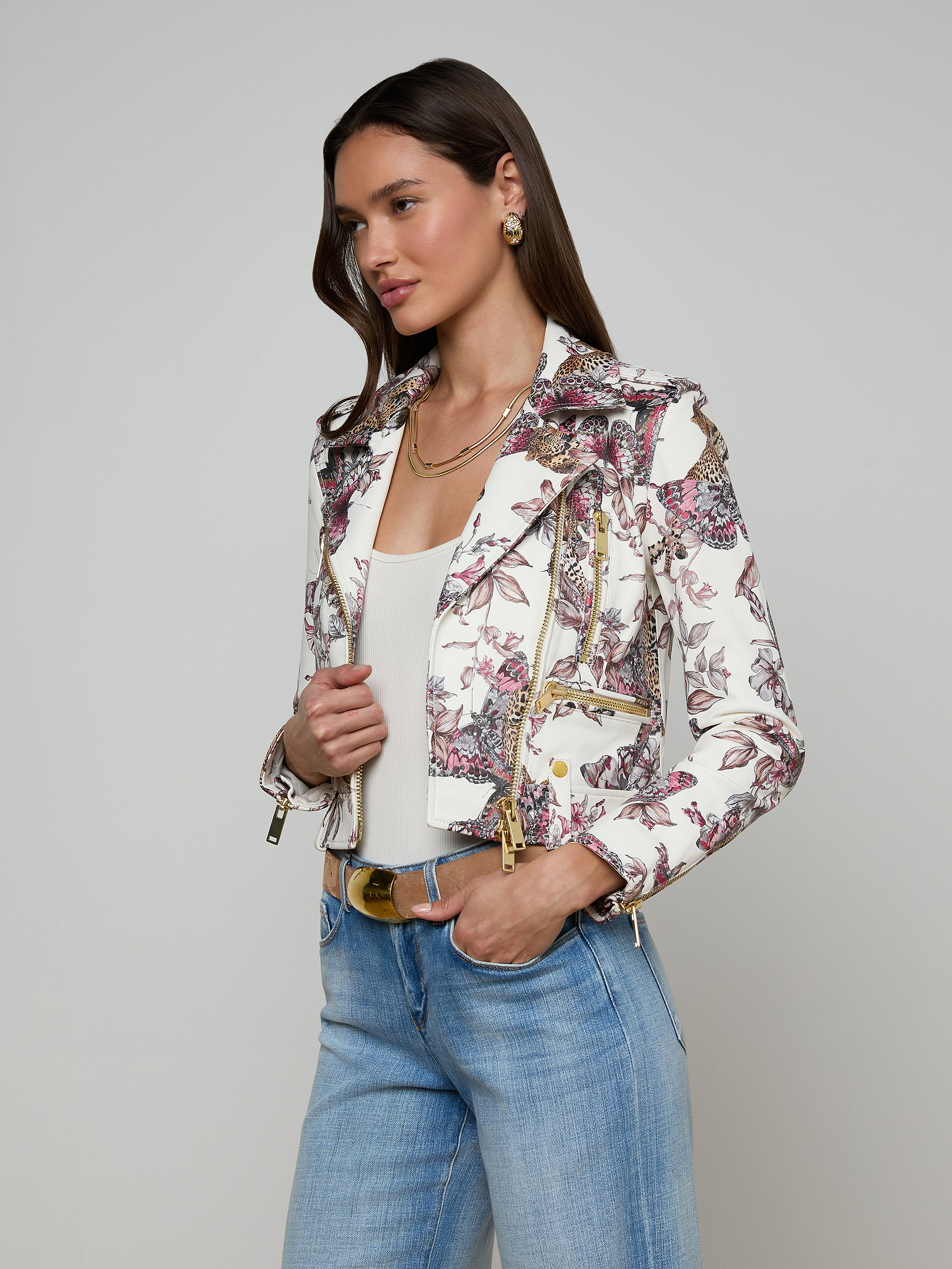 A woman wears the Blakely Leather Biker Jacket, featuring a white base and butterfly jungle print, layered over a white top with light blue jeans and accented by gold jewelry, all set against a plain light background.