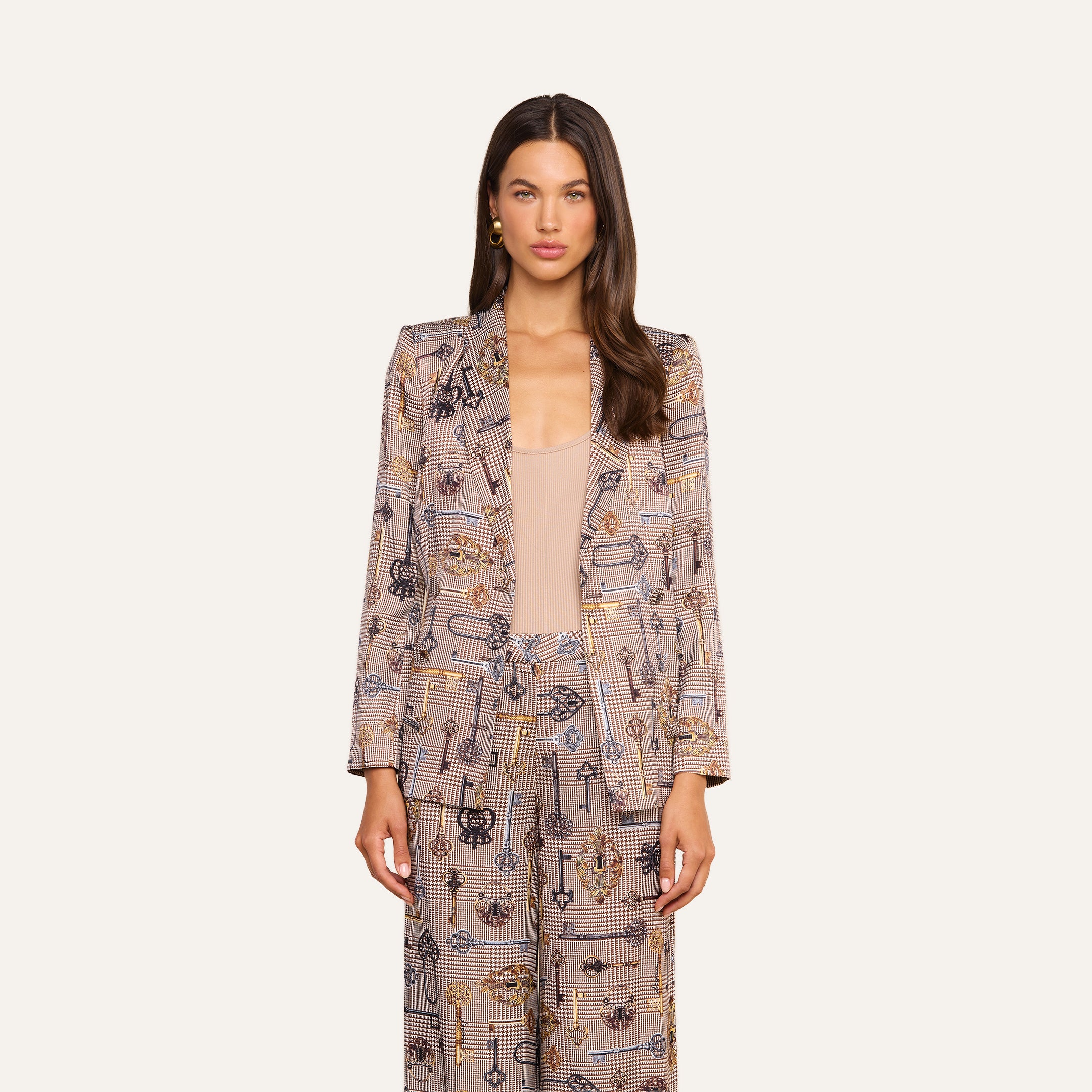 A woman with long brown hair stands against a plain off-white background, facing forward with a neutral expression and her arms relaxed at her sides. She wears the Aven Wrap Blazer in a brown houndstooth pattern layered over a beige top, creating a modern suiting ensemble.