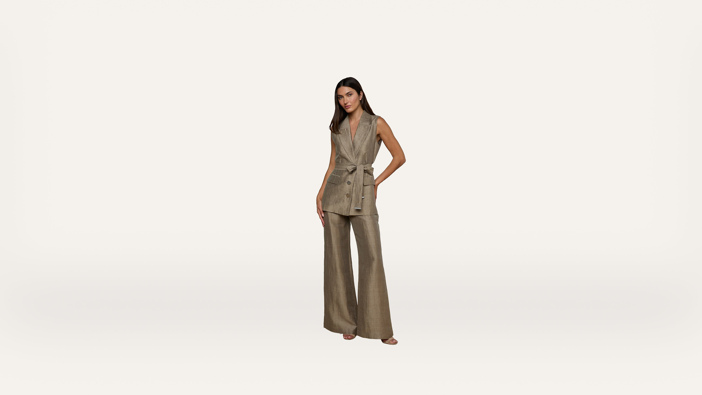 Woman standing against a plain white background, wearing a taupe sleeveless belted vest and wide-leg trousers. One hand is in her pocket, and she faces forward with a neutral expression.