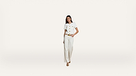 A woman wearing a white short-sleeved top with gold buttons and white high-waisted trousers stands against a plain white background. She has one hand in her pocket and is wearing beige heels.