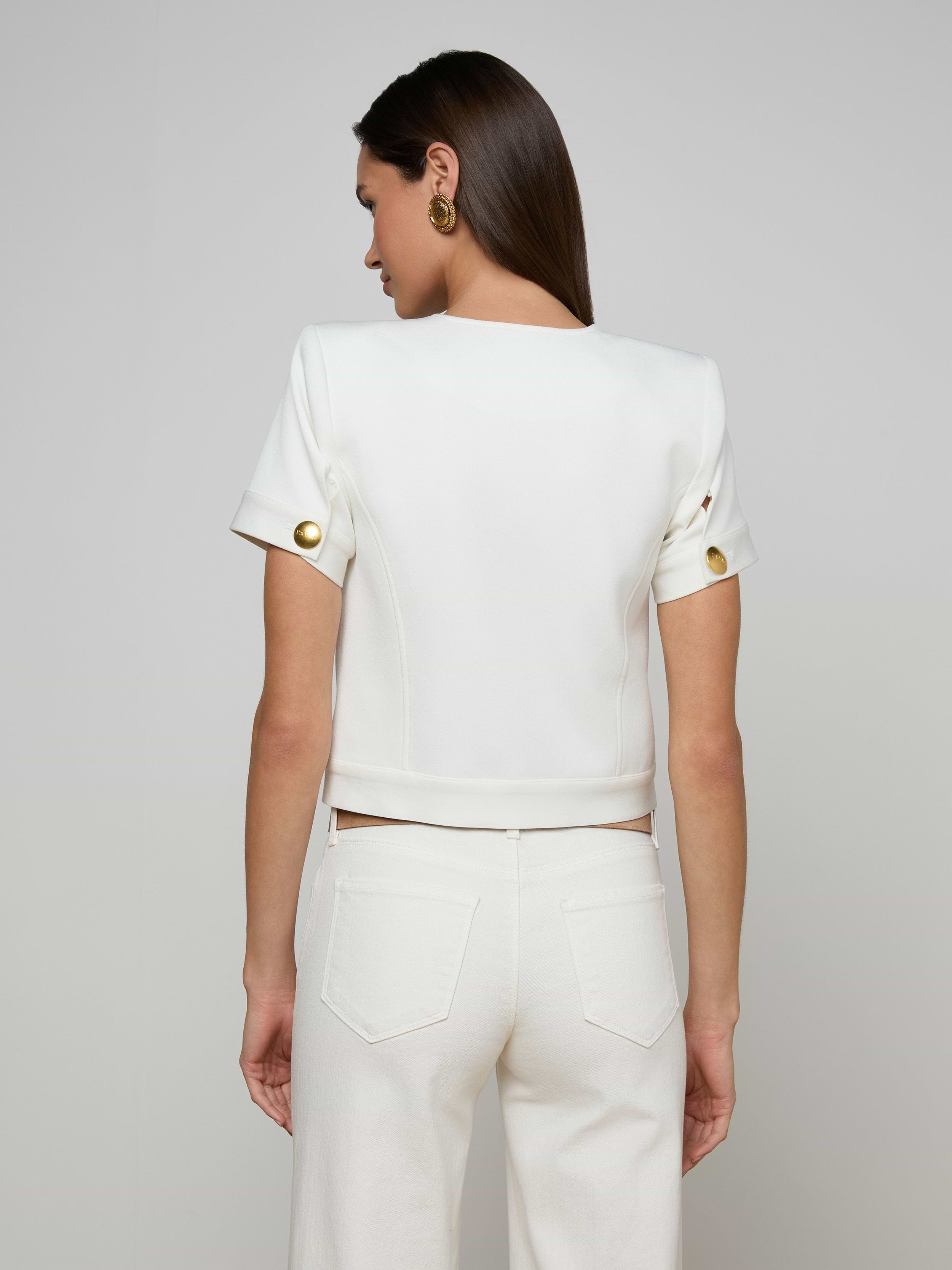 A woman stands with her back to the camera, wearing the Nawa Shirt Jacket—a tailored, collarless white piece with gold buttons on the sleeves—paired with matching white pants. Her straight brown hair and gold earrings complement the ensemble against a plain light background.