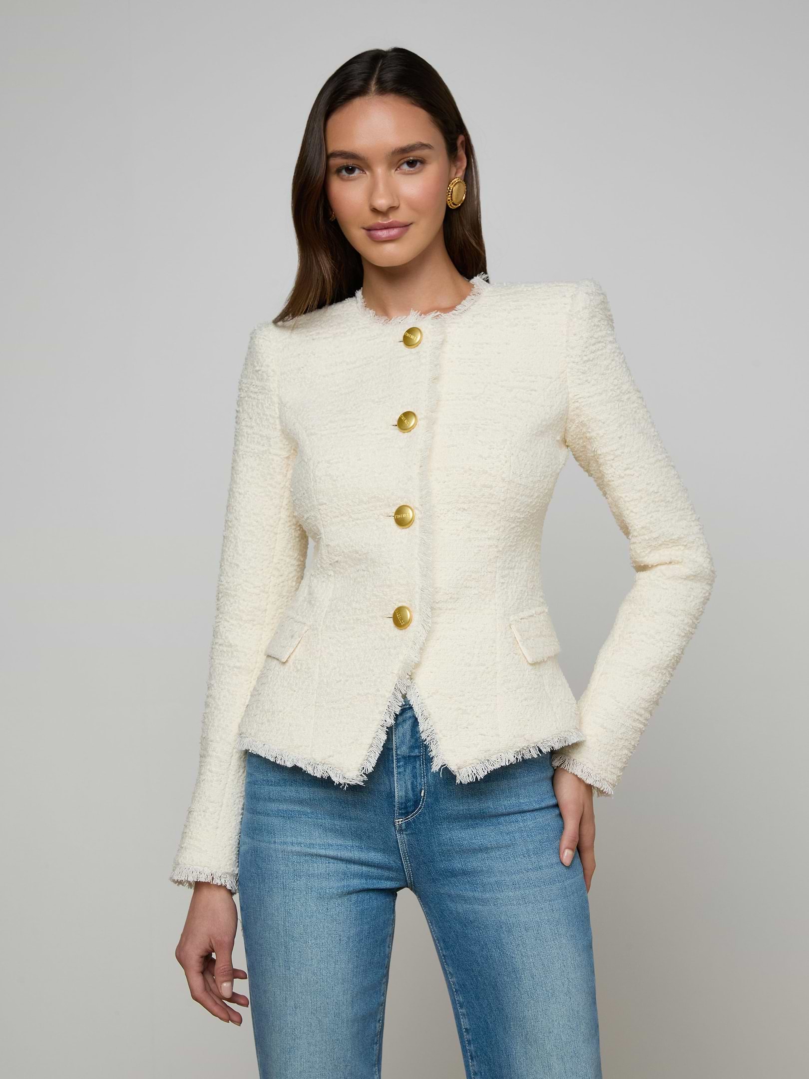 A woman wears the Nerina Tweed Blazer—a fitted, textured, white collarless jacket with gold buttons—paired with blue jeans, standing against a plain light background.