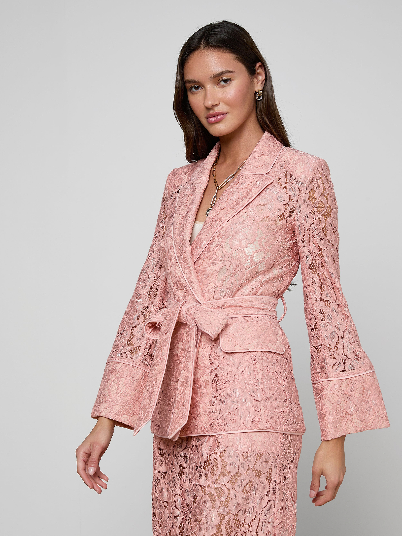 A woman stands against a plain, light background, facing slightly to the side with a neutral expression, wearing the Jordana Lace Blazer—a pink sheer floral lace suit featuring a belted blazer and matching skirt.