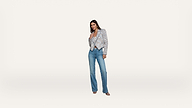 A woman stands against a plain white background, wearing a patterned blazer, a light top, blue jeans, and open-toed heels, with one hand touching her chest and the other by her side.