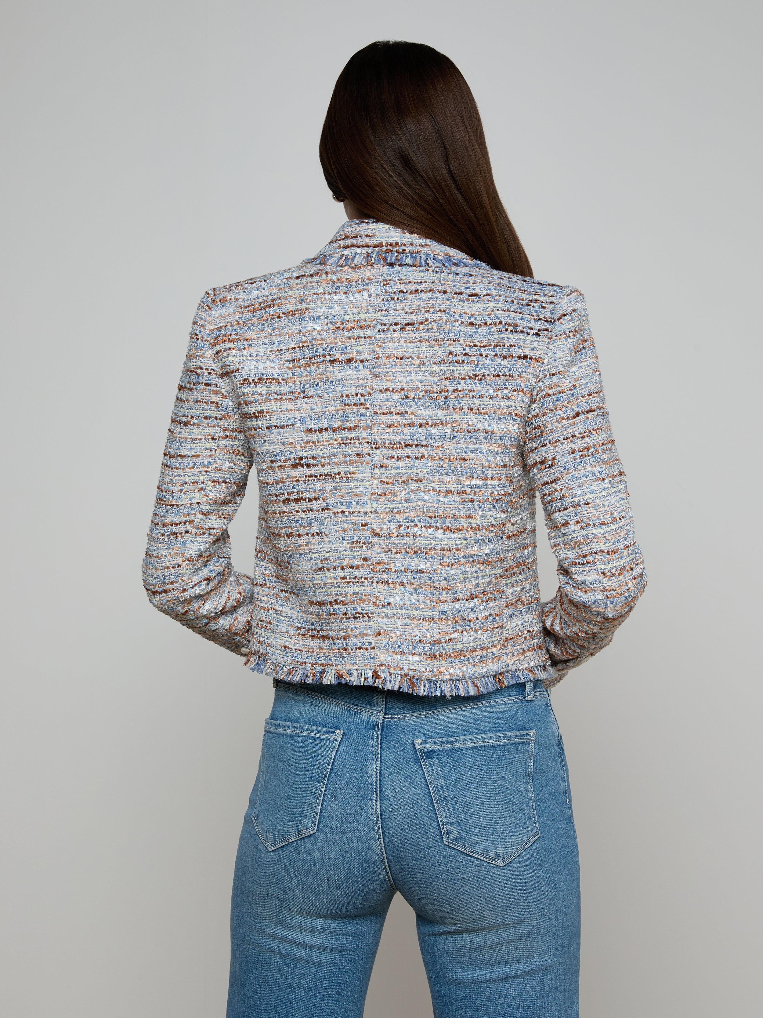 A woman with long brown hair, seen from the back, is wearing the Marv Tweed Blazer—a textured, multi-colored design with statement buttons—paired with high-waisted blue jeans against a plain light background.