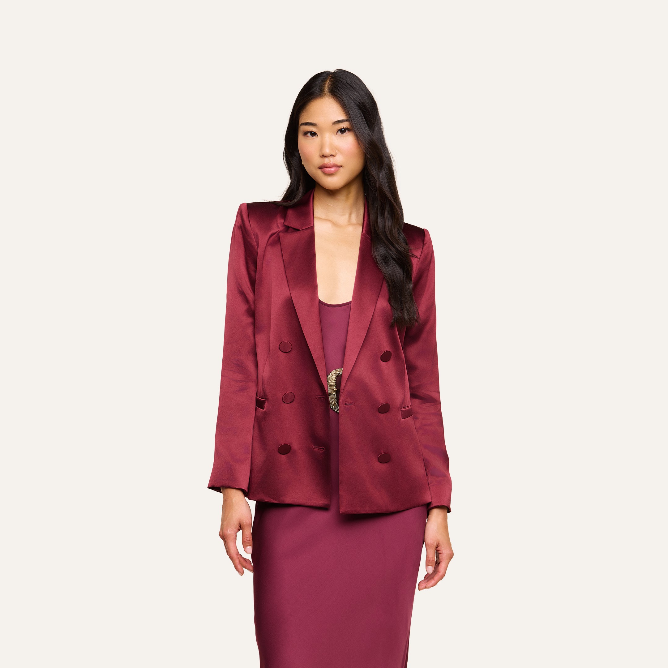 A woman with long dark hair wears the Sena Silk Blazer in merlot red over a matching maroon dress, standing against a plain, light background.