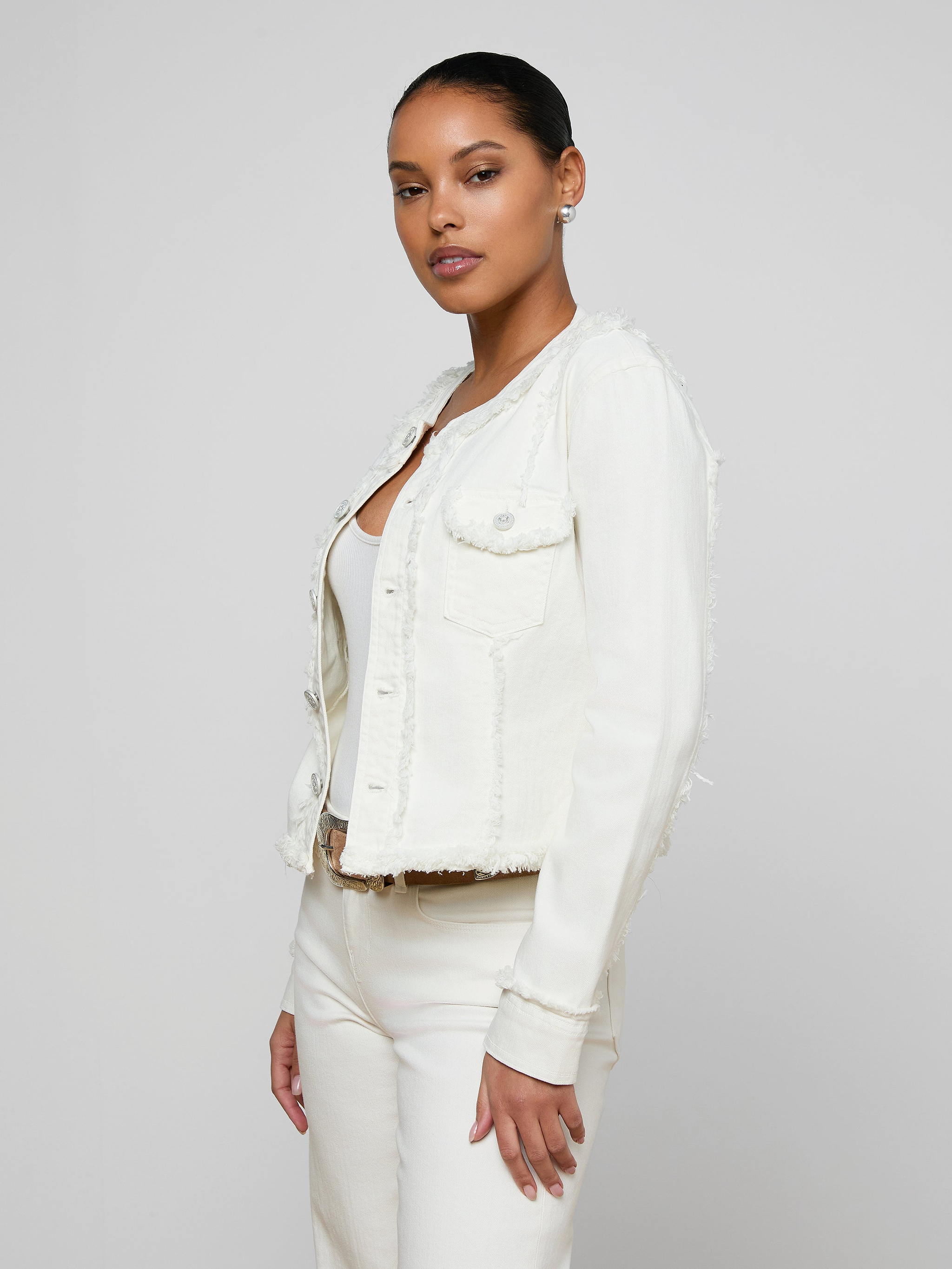 A woman stands against a plain background wearing the Yarin Collarless Fray Jacket—crafted from stretch denim without a collar—layered over a white top and white pants, looking confidently at the camera with her hands relaxed by her sides.