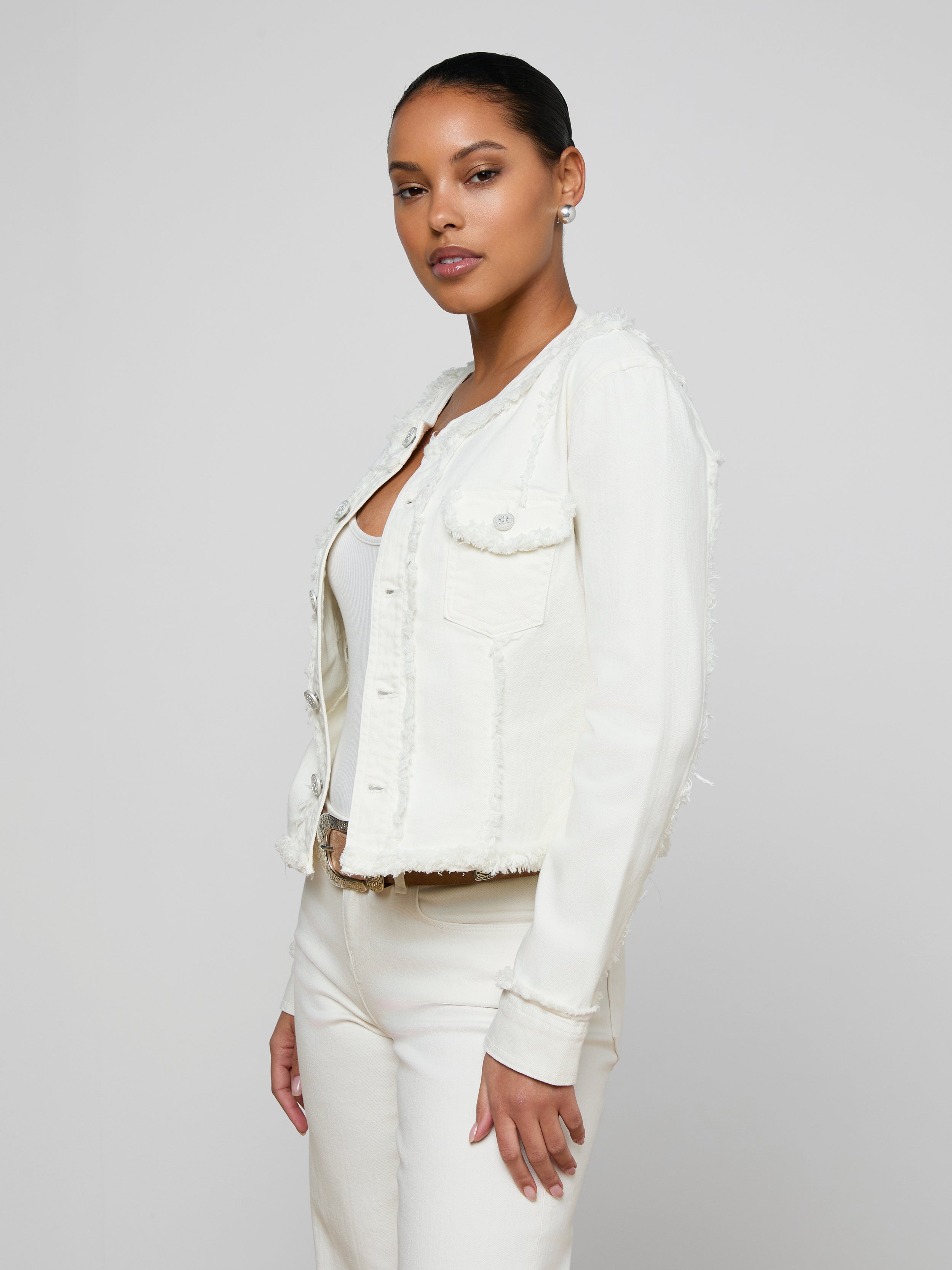 A woman stands against a plain background wearing the Yarin Collarless Fray Jacket—crafted from stretch denim without a collar—layered over a white top and white pants, looking confidently at the camera with her hands relaxed by her sides.