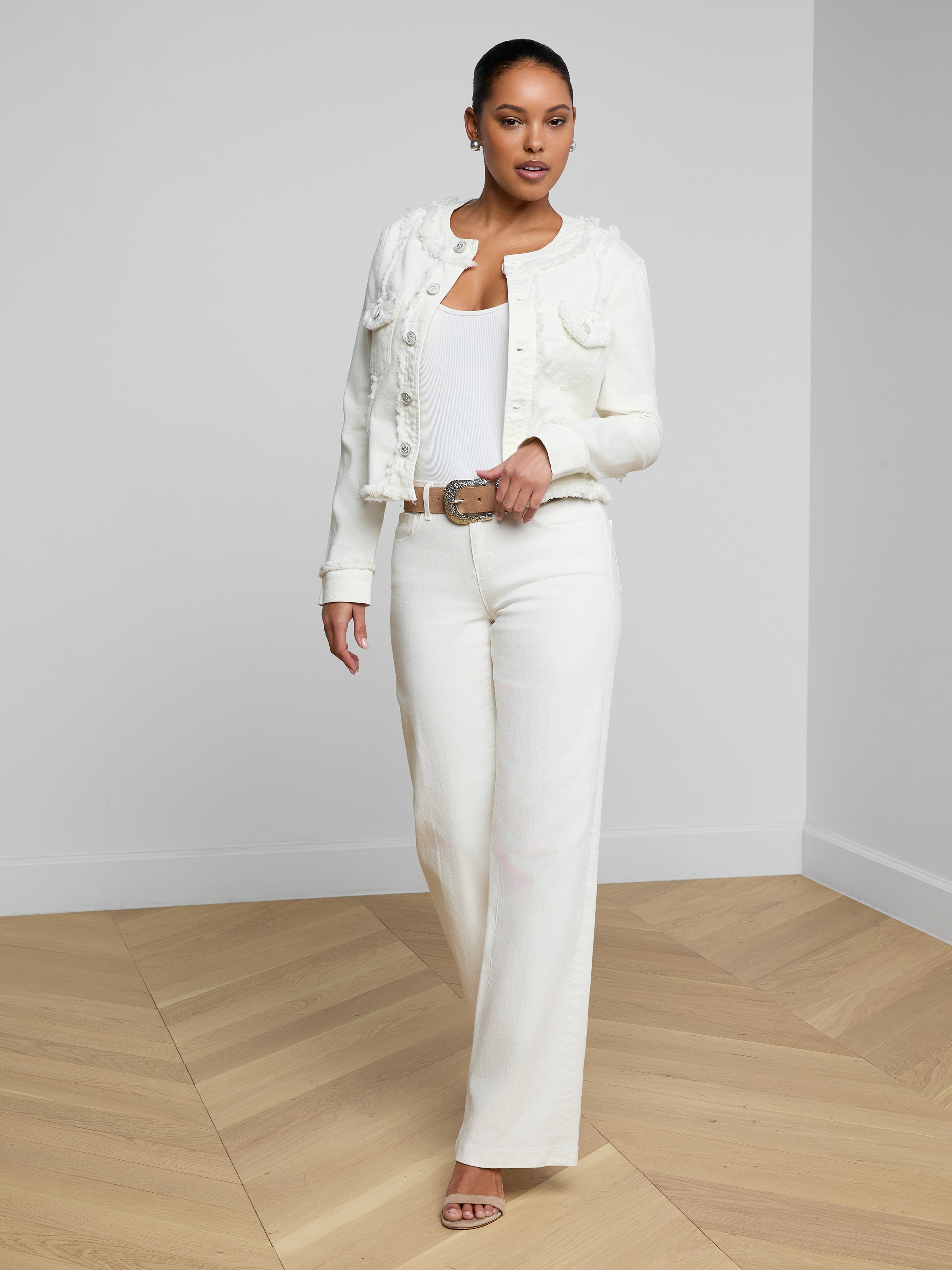 A woman stands confidently against a white background, wearing the Yarin Collarless Fray Jacket over a white top and wide-leg white pants with beige heels.