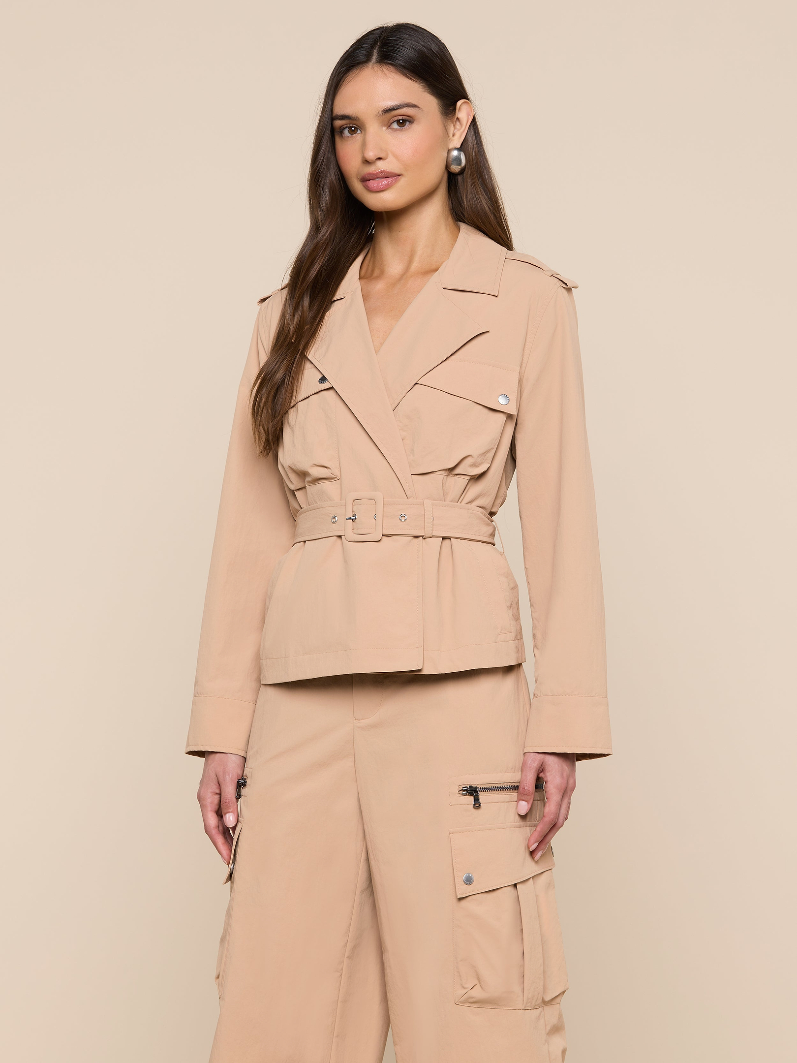 A woman stands against a plain light background, wearing the Calida Windbreaker Trench—a belted, beige utility jacket crafted from water-repellent nylon—paired with matching cargo pants. Her hair is down and her hands rest by her sides.