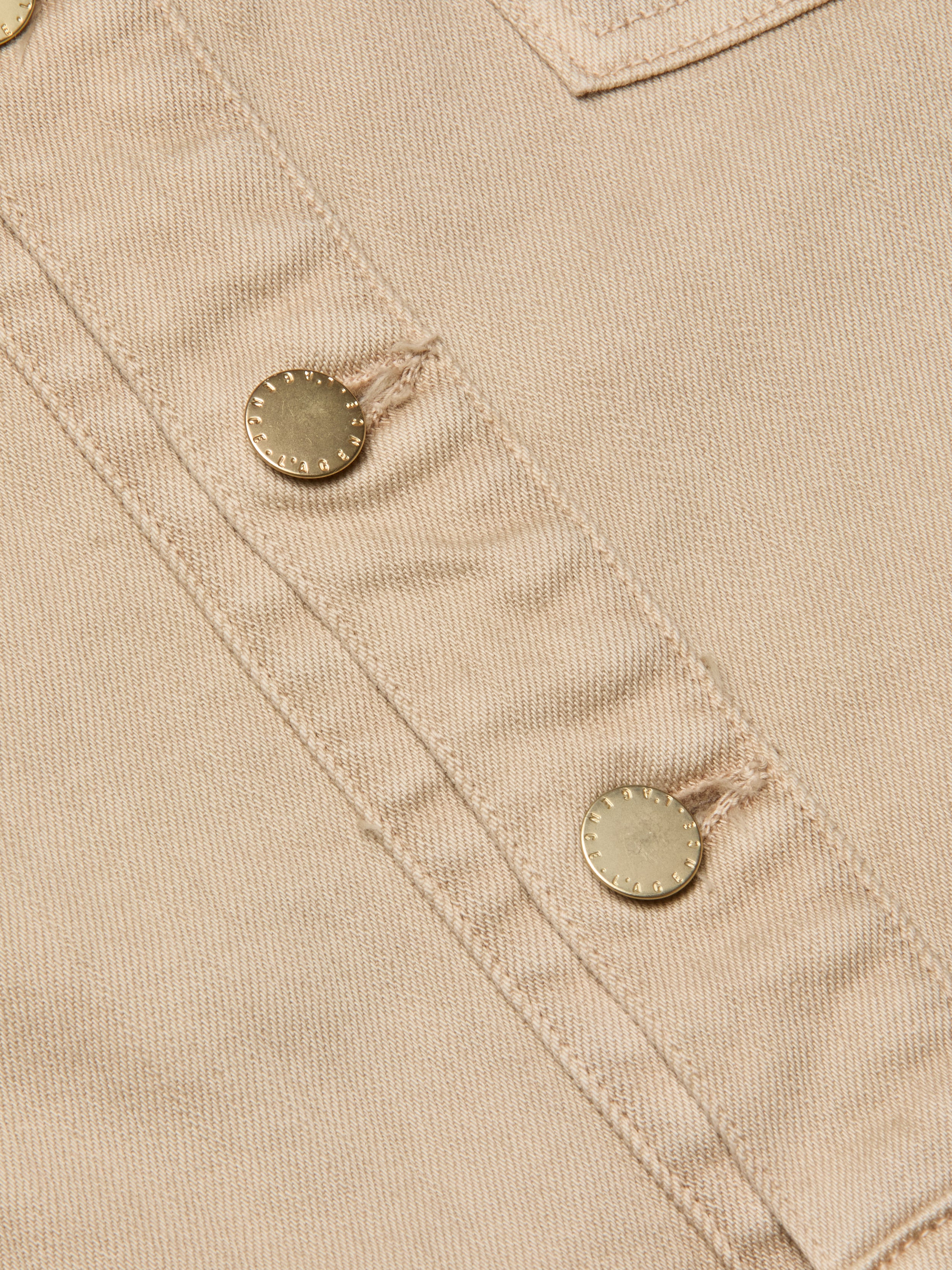 Close-up of beige fabric featuring two metallic buttons sewn in a diagonal line, highlighting the visible stitching and textured detailing characteristic of the Kirstie Jean Shirt Jacket.