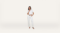 A woman stands against a plain white background, wearing a short-sleeved white button-up shirt, white high-waisted pants, and heeled sandals. She has one hand on her hip and the other touching her chin.