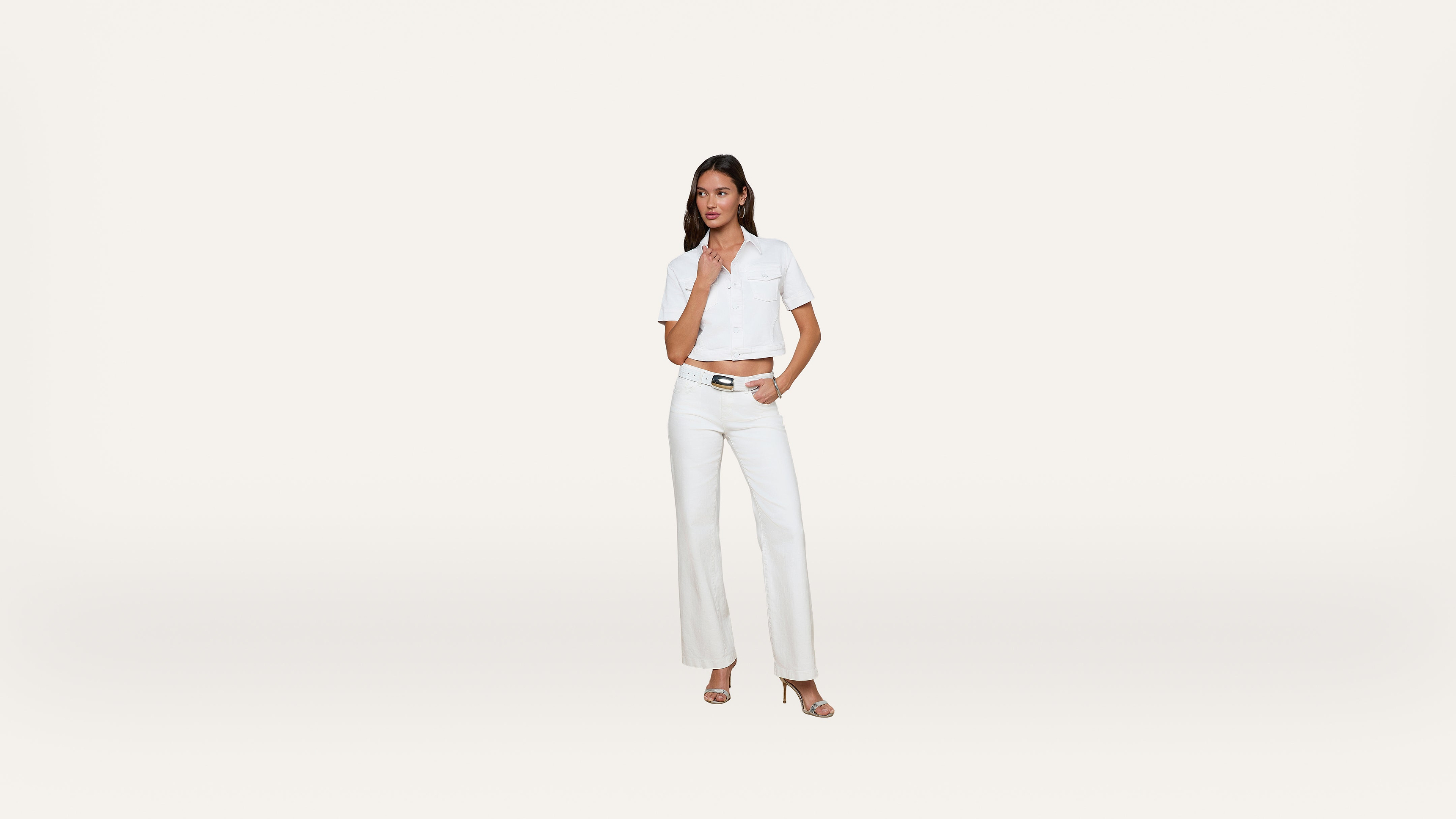 A woman stands against a plain white background, wearing a short-sleeved white button-up shirt, white high-waisted pants, and heeled sandals. She has one hand on her hip and the other touching her chin.