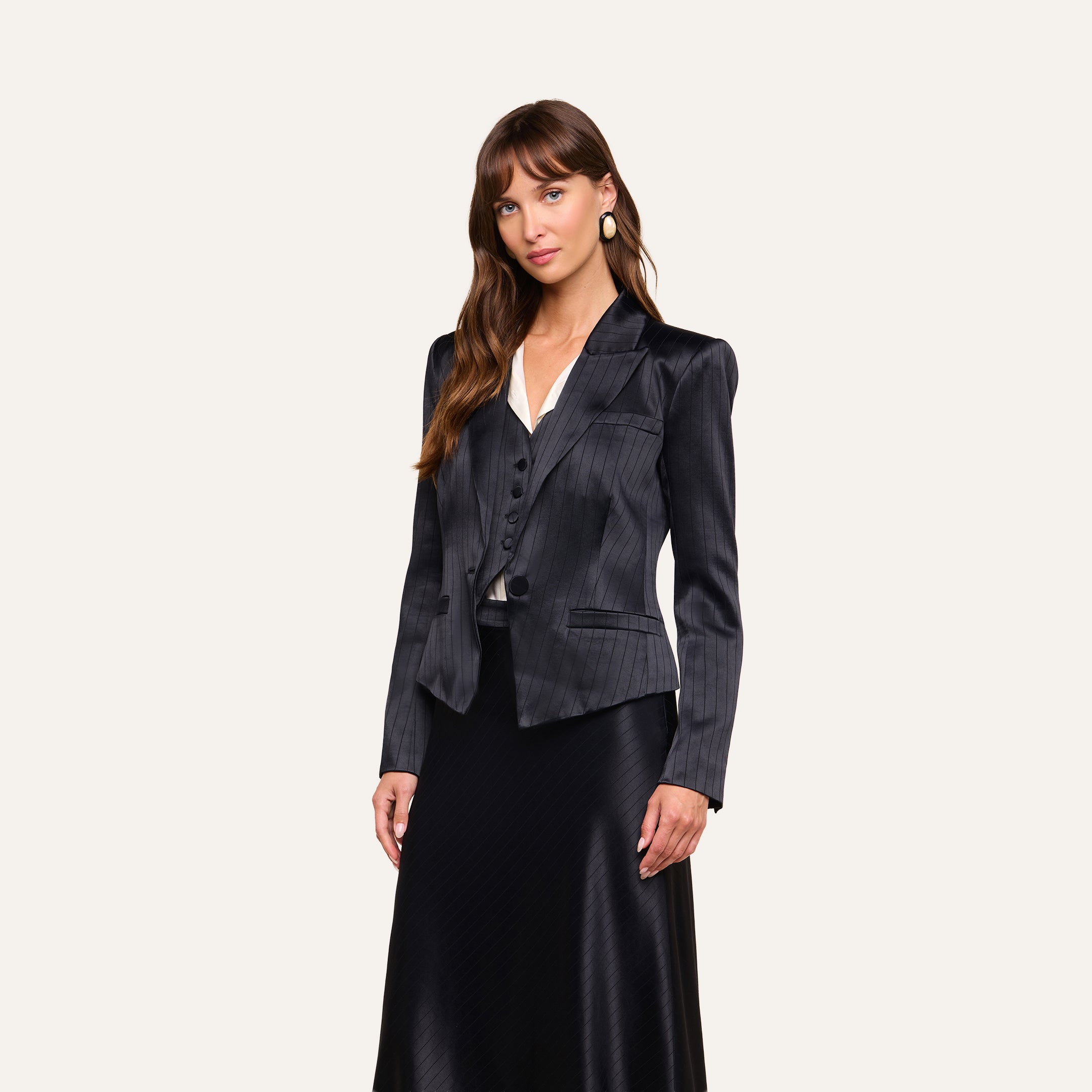 A woman stands against a plain background, wearing the black pinstripe Misa Blazer with a matching vest and a long black skirt. Her ensemble features elements of satin suiting and is complemented by a crisp white top. She has long brown hair with bangs and is looking at the camera.