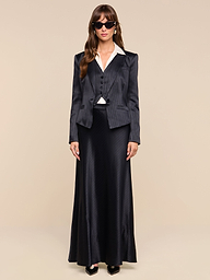 A woman wearing sunglasses and the Misa Blazer over a white shirt, paired with a black vest and a long black skirt, stands against a plain white background.