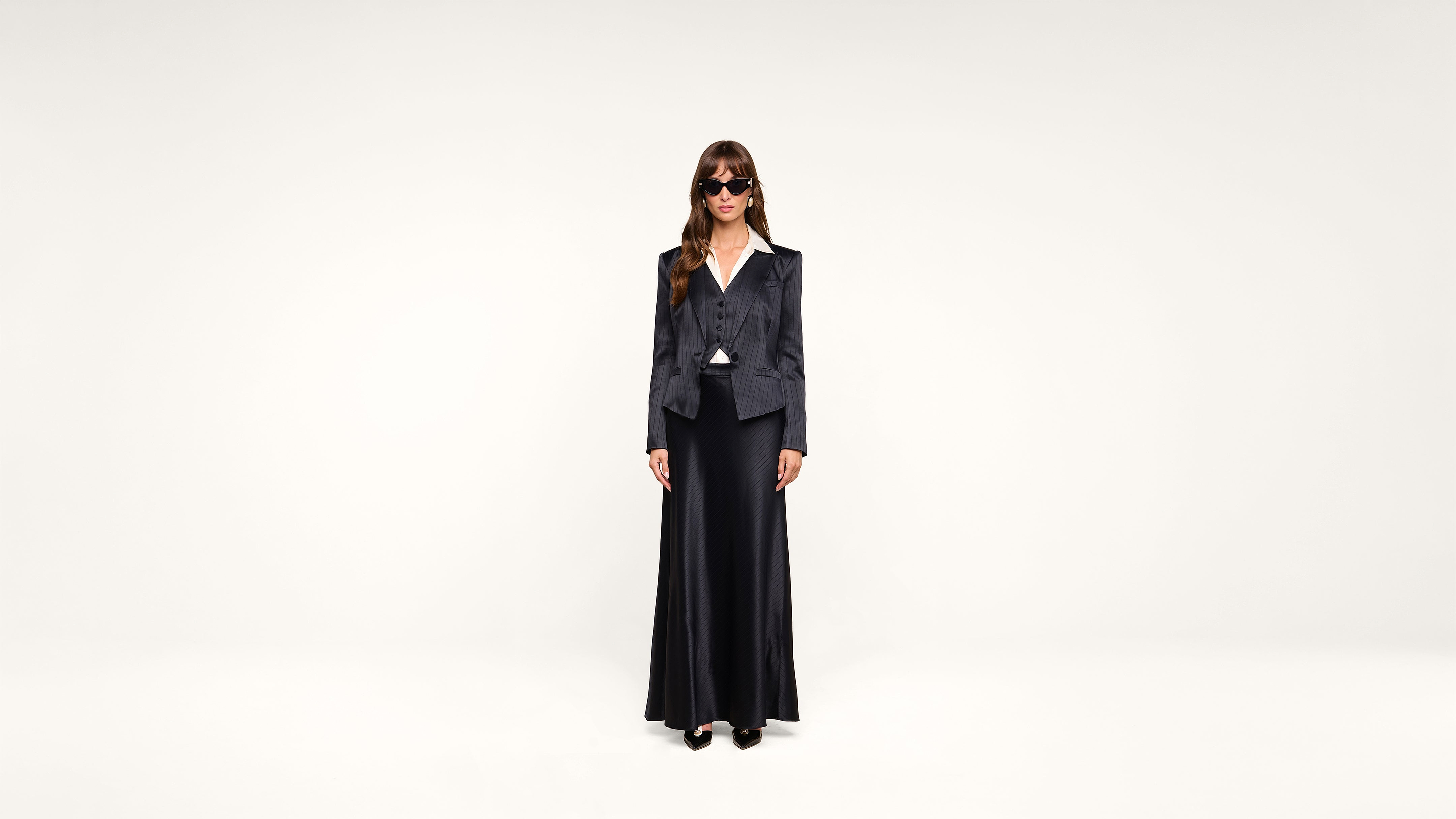 A woman stands against a plain white background wearing sunglasses, a dark blazer, a matching long skirt, and black shoes.